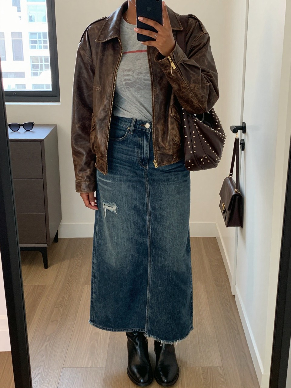 Mirror selfie of a Black woman wearing a dark indigo distressed denim maxi skirt with a front slit, a faded grey fitted graphic T-shirt, a worn chocolate-brown leather jacket, black leather ankle boots, and a dark brown studded shoulder bag, clearly showing the exact clothing items, colors, fabrics, and accessories listed, holding a phone in front of a stylish city apartment mirror, cropped at the neck so the face does not show, relaxed casual selfie pose, natural window light, sunglasses placed on a dresser and a handbag hanging on a door handle, natural uncluttered setting, latest iPhone photo quality.