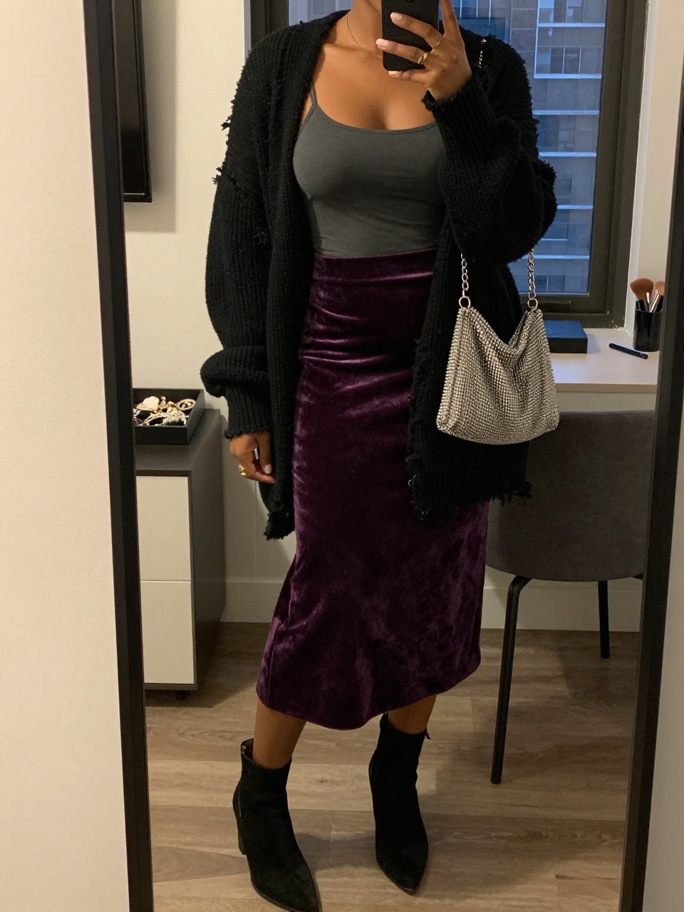 Mirror selfie of a Black woman wearing a deep purple velvet midi skirt, a fitted charcoal-grey camisole, an oversized distressed black cardigan, black suede ankle boots, and a silver chainmail mini bag, clearly showing the exact clothing items, colors, fabrics, and accessories listed, holding a phone in front of a stylish city apartment mirror, cropped at the neck so the face does not show, relaxed casual selfie pose, warm moody lighting, a jewelry tray on the vanity and a makeup brush on the counter, natural evening setting, latest iPhone photo quality.