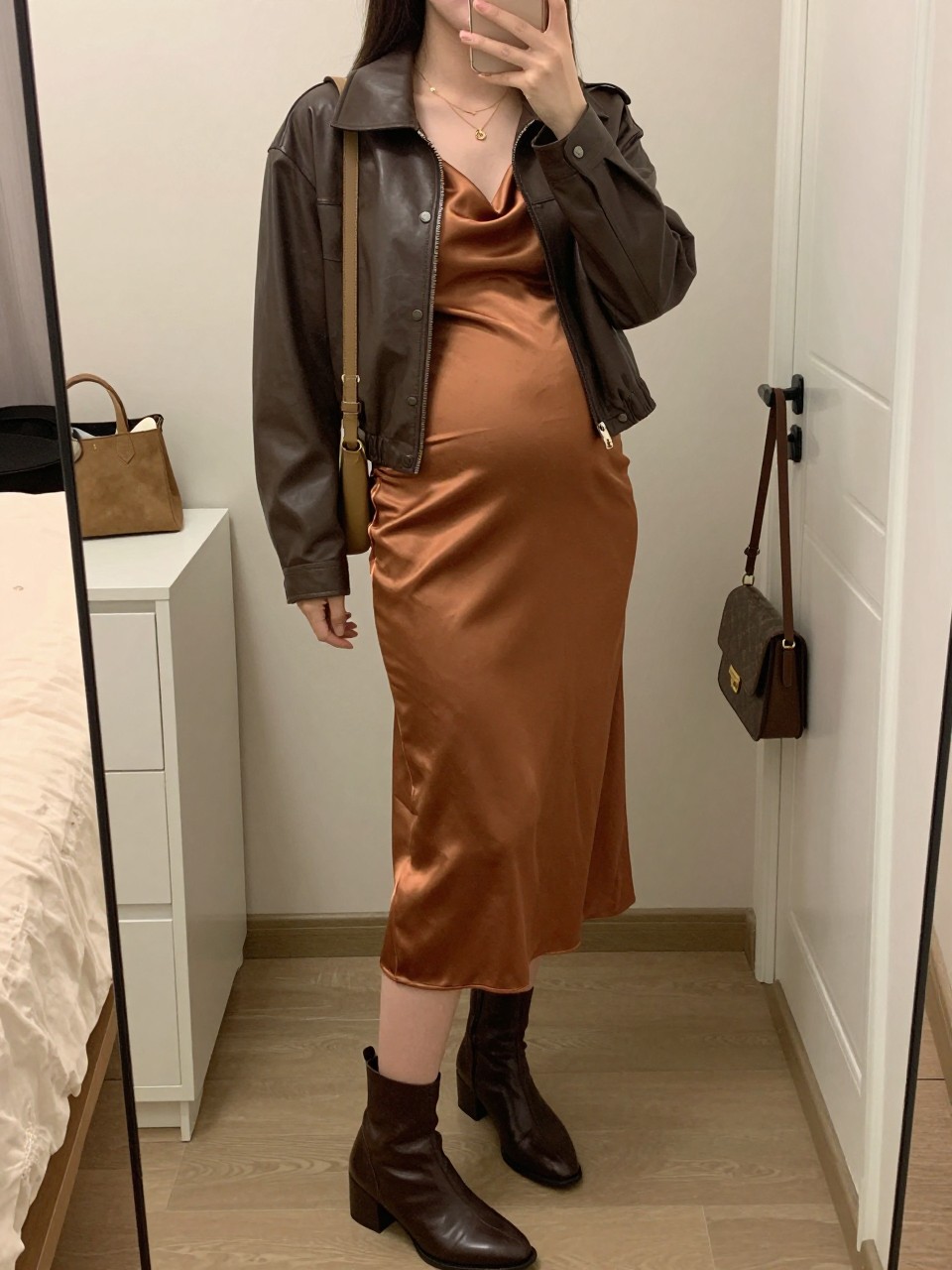 Mirror selfie of a pregnant woman wearing a copper satin maternity midi dress with a cowl neckline and softly fitted waist, a cropped chocolate-brown leather jacket, dark brown heeled ankle boots, a small tan suede shoulder bag, and delicate gold layered necklaces, clearly showing the exact clothing items, colors, fabrics, and accessories listed, holding a phone in front of a feminine-themed bedroom mirror, cropped at the neck so the face does not show, with a relaxed casual selfie pose, warm moody lighting, a cosmetic bag on the counter and a handbag hanging on a door handle, realistic and uncluttered, latest iPhone photo quality.