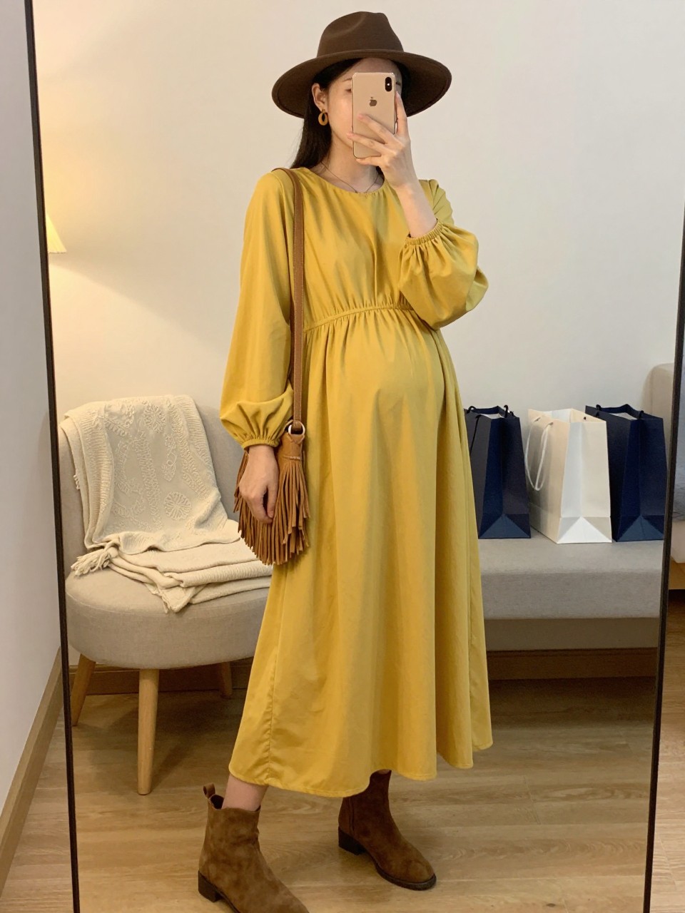 Mirror selfie of a pregnant woman wearing a flowing mustard-yellow maternity maxi dress with long balloon sleeves and a gathered empire waist, brown suede ankle boots, a chocolate-brown felt wide-brim hat, a tan fringed crossbody bag, and small wooden hoop earrings, clearly showing the exact clothing items, colors, fabrics, and accessories listed, holding a phone in front of a cozy bedroom mirror setup, cropped at the neck so the face does not show, with a relaxed casual selfie pose, evening golden-hour lighting, a folded blanket on a chair and shopping bags beside a bench, natural and casually styled, latest iPhone photo quality.