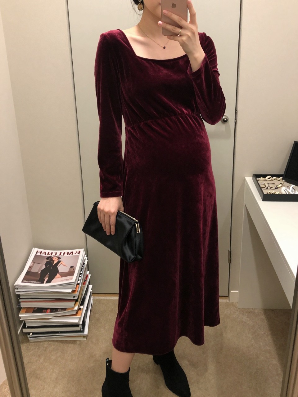 Mirror selfie of a pregnant woman wearing a deep burgundy velvet maternity maxi dress with a square neckline, long fitted sleeves, and a softly gathered waist, black suede ankle boots, a small black satin clutch, and antique-gold pendant earrings, clearly showing the exact clothing items, colors, fabrics, and accessories listed, holding a phone in front of a minimal dressing room mirror, cropped at the neck so the face does not show, with a relaxed casual selfie pose, warm moody lighting, a stack of fashion magazines on a side table and a jewelry tray on the vanity, elegant but natural, latest iPhone photo quality.