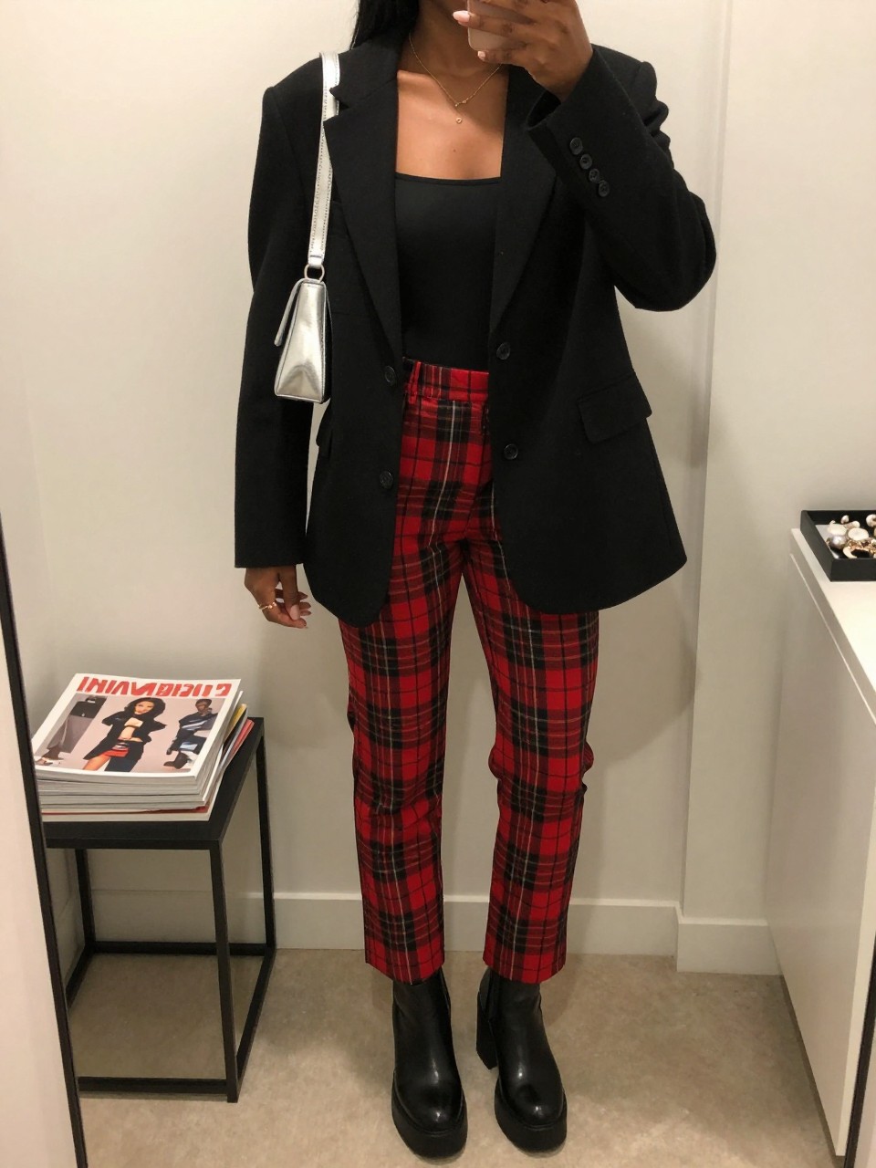 Mirror selfie of a Black woman wearing red and black tartan straight-leg trousers, a fitted black square-neck bodysuit, an oversized black wool blazer, black leather platform ankle boots, and a silver metallic shoulder bag, clearly showing the exact clothing items, colors, fabrics, and accessories listed, holding a phone in front of a minimal dressing room mirror, cropped at the neck so the face does not show, relaxed casual selfie pose, soft indoor lighting, a stack of fashion magazines on a side table and a jewelry tray on the vanity, polished but natural, latest iPhone photo quality.