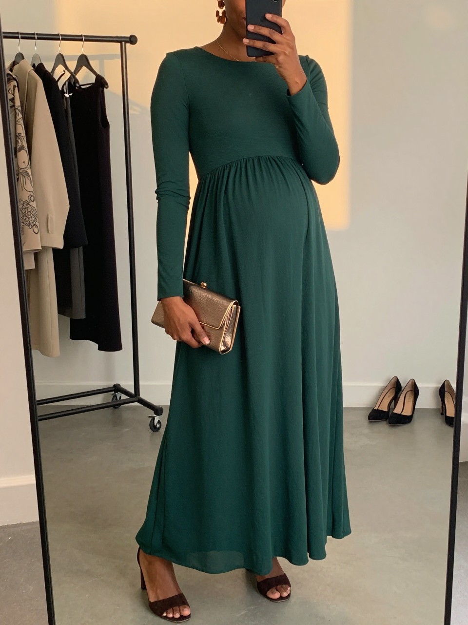 Mirror selfie of a Black pregnant woman wearing a floor-length forest-green maternity gown with a fitted bodice, long sleeves, and a full flowing skirt, dark brown suede block heels, bronze statement earrings, and a small metallic bronze clutch, clearly showing the exact clothing items, colors, fabrics, and accessories listed, holding a phone in front of a fashion studio mirror corner, cropped at the neck so the face does not show, with a relaxed casual selfie pose, evening golden-hour lighting, a clothing rack with garments behind and a pair of heels on the floor nearby, natural editorial setting, latest iPhone photo quality.