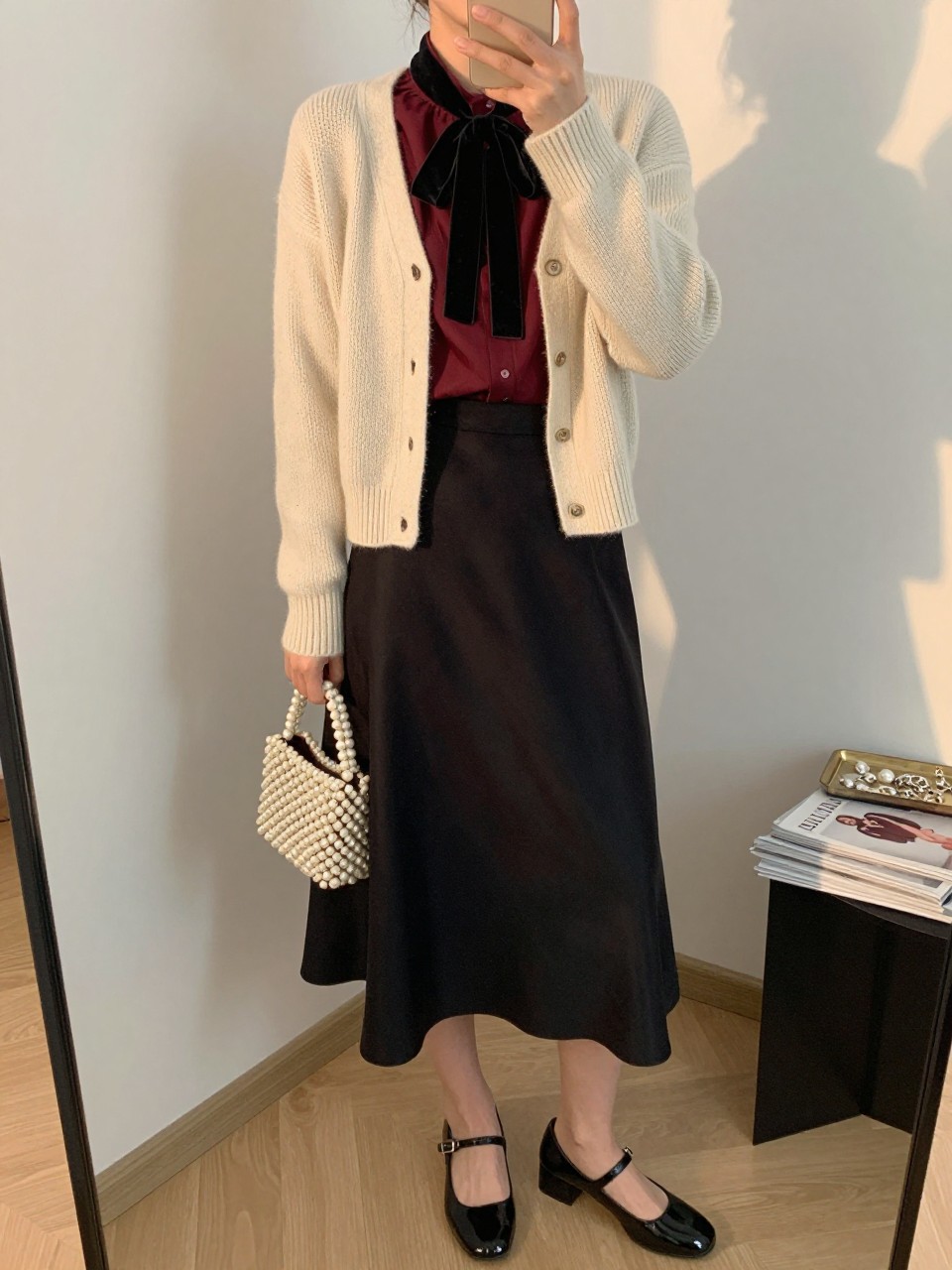 Image Prompt mirror selfie of a woman wearing an open cream fitted fine-knit cardigan over a burgundy chiffon blouse with a black velvet neck bow tucked into a black satin A-line midi skirt, black patent leather Mary Jane heels, and a cream beaded mini handbag, clearly showing the exact clothing items, colors, fabrics, bow, shoes, and bag listed in the Outfit Pieces section, holding a phone in front of a mirror, cropped at NECK so the face does not show, relaxed polished selfie pose in a fashion studio mirror corner, evening golden hour light, with a jewelry tray on a nearby surface and a stack of fashion magazines on a side table, realistic knit, chiffon, velvet, satin, patent leather, and beadwork textures, latest iPhone photo quality