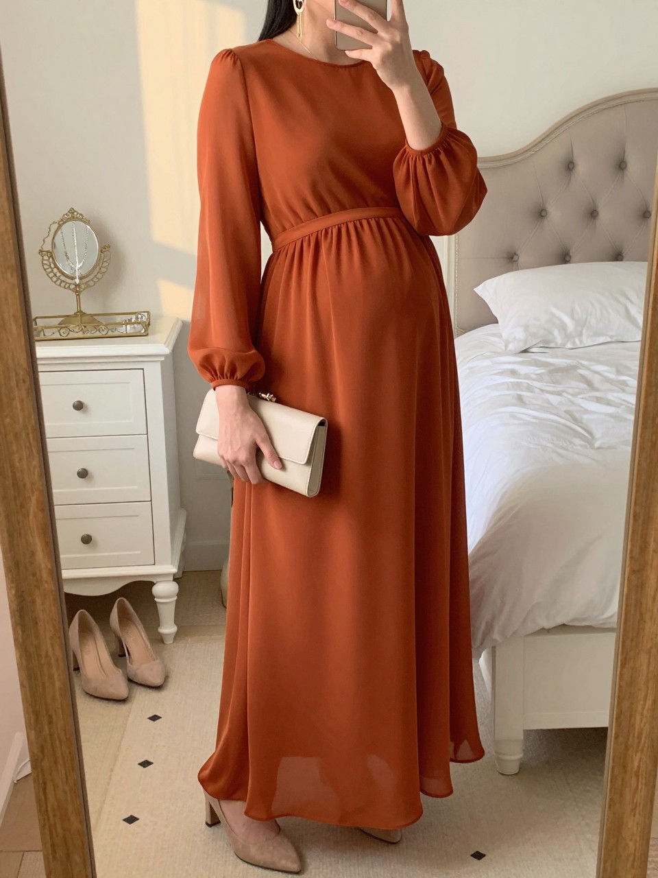 Mirror selfie of a pregnant woman wearing a floor-length rust-orange chiffon maternity gown with long sheer sleeves and a fitted empire waist, nude suede block heels, delicate gold drop earrings, and a small cream clutch, clearly showing the exact clothing items, colors, fabrics, and accessories listed, holding a phone in front of a feminine-themed bedroom mirror, cropped at the neck so the face does not show, with a relaxed casual selfie pose, evening golden-hour lighting, a jewelry tray on the vanity and a pair of heels on the floor nearby, natural and uncluttered, latest iPhone photo quality.