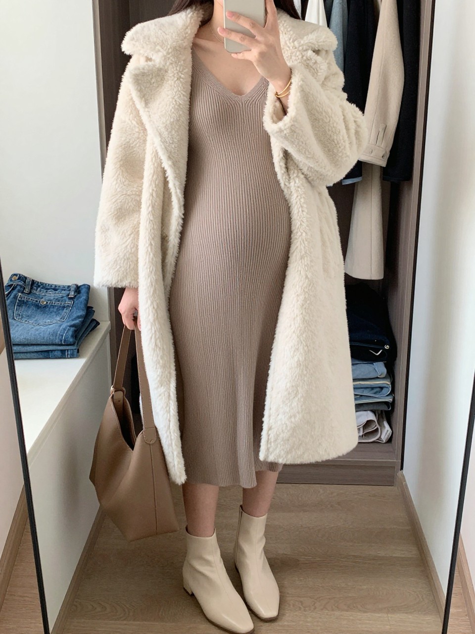 Mirror selfie of a pregnant woman wearing a fitted mocha ribbed-knit maternity midi dress, an open ivory teddy-fleece coat, cream leather ankle boots, a soft brown hobo bag, and thin gold bangles, clearly showing the exact clothing items, colors, fabrics, and accessories listed, holding a phone in front of a walk-in closet mirror, cropped at the neck so the face does not show, with a relaxed casual selfie pose, natural window light, an open closet with clothes visible and folded denim jeans on a bench, cozy and realistic, latest iPhone photo quality.