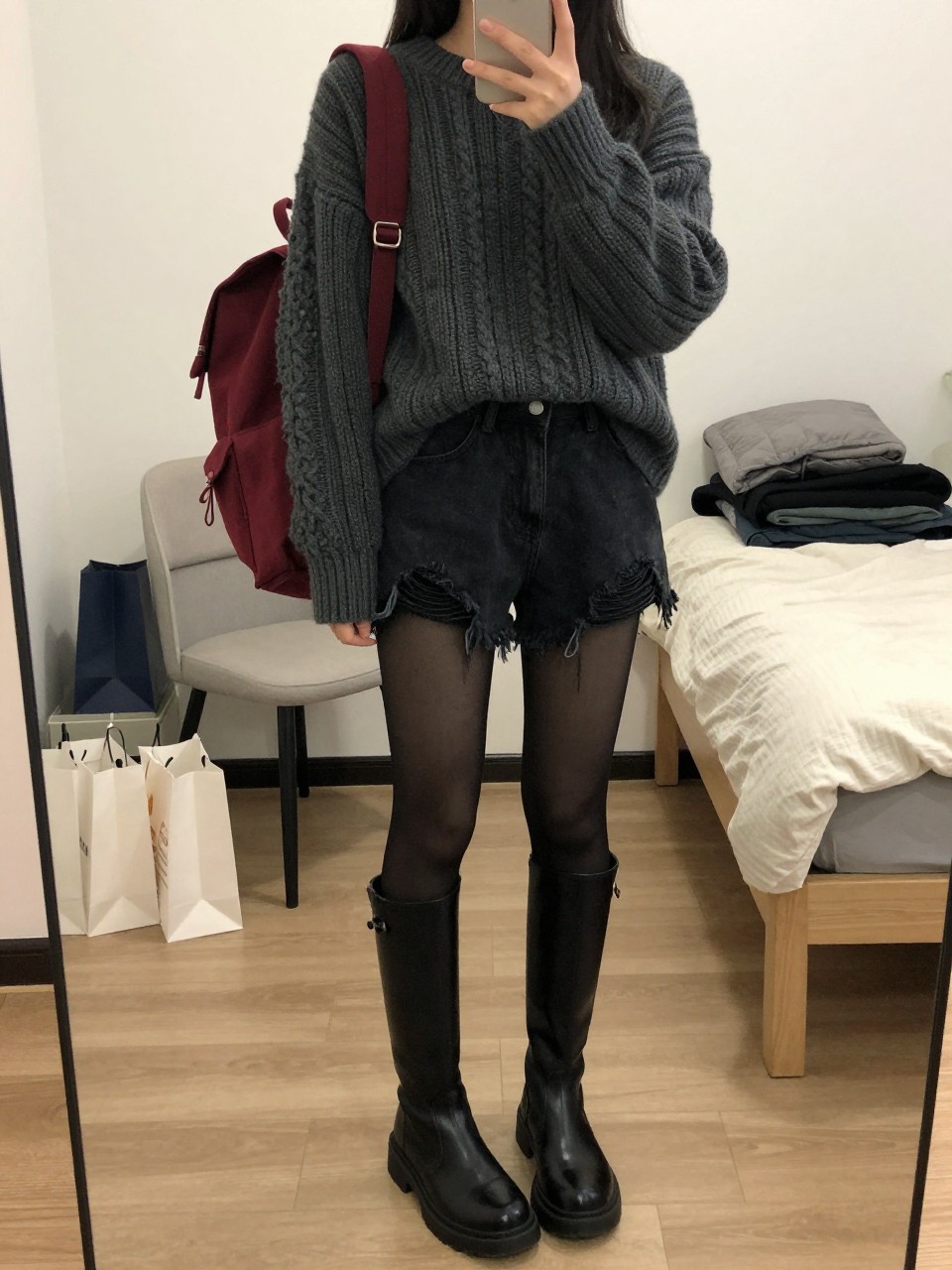 Mirror selfie of a woman wearing distressed black denim shorts, an oversized dark grey cable-knit sweater, opaque black tights, knee-high black leather combat boots, and a burgundy canvas backpack, clearly showing the exact clothing items, colors, fabrics, and accessories listed, holding a phone in front of a cozy bedroom mirror setup, cropped at the neck so the face does not show, relaxed casual selfie pose, soft indoor lighting, shopping bags beside a chair and a folded blanket on a bench, realistic and uncluttered, latest iPhone photo quality.