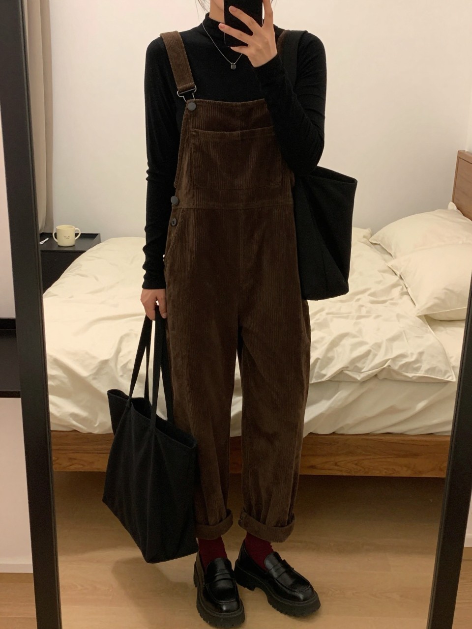 Mirror selfie of a woman wearing loose dark brown corduroy overalls, a fitted black ribbed mock-neck top, black platform loafers, burgundy crew socks, and a black canvas tote bag, clearly showing the exact clothing items, colors, fabrics, and accessories listed, holding a phone in front of a cozy bedroom mirror setup, cropped at the neck so the face does not show, relaxed casual selfie pose, warm moody lighting, soft pillows on an unmade bed and a coffee mug on a bedside table, believable lived-in environment, latest iPhone photo quality.