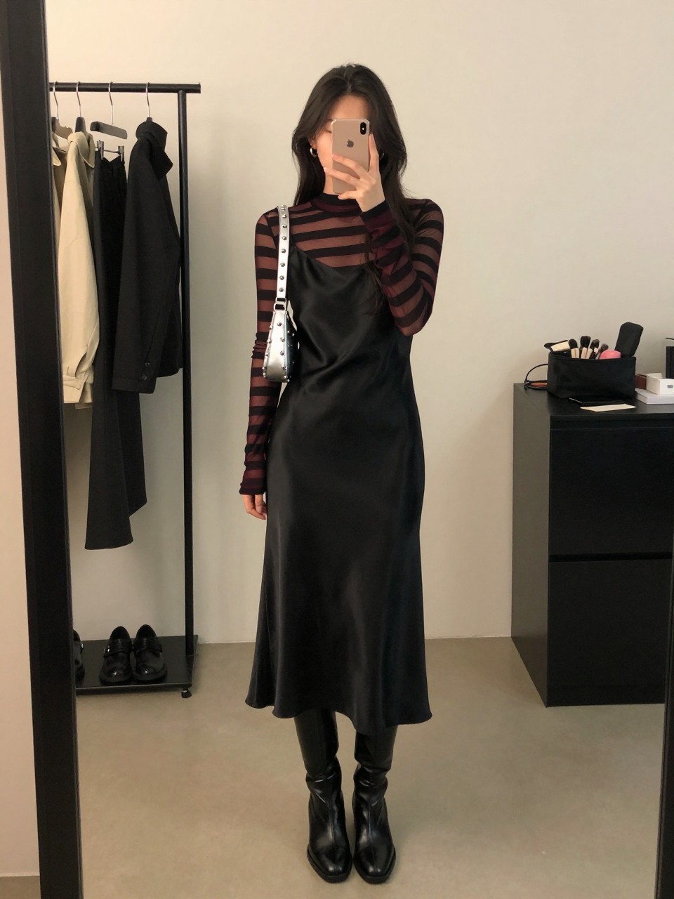 Mirror selfie of a woman wearing a black satin midi slip dress layered over a fitted black and burgundy striped mesh long-sleeve top, black leather knee-high boots, a silver-studded mini shoulder bag, and small silver hoop earrings, clearly showing the exact clothing items, colors, fabrics, and accessories listed, holding a phone in front of a fashion studio mirror corner, cropped at the neck so the face does not show, relaxed casual selfie pose, warm moody lighting, a clothing rack with garments behind and a makeup bag on the counter, natural and unstaged, latest iPhone photo quality.
