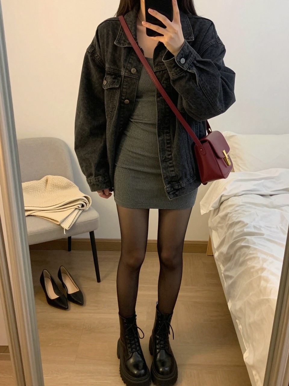 Mirror selfie of a woman wearing a fitted charcoal-grey ribbed mini dress, an oversized faded black denim jacket, sheer black tights, chunky black lace-up boots, and an oxblood leather crossbody bag, clearly showing the exact clothing items, colors, fabrics, and accessories listed, holding a phone in front of a feminine-themed bedroom mirror, cropped at the neck so the face does not show, relaxed casual selfie pose, evening golden-hour lighting, a folded blanket on a chair and a pair of heels on the floor nearby, natural and uncluttered, latest iPhone photo quality.