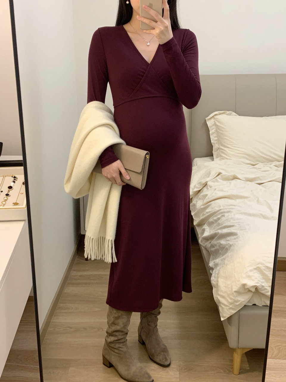 Mirror selfie of a pregnant woman wearing a deep plum long-sleeve maternity wrap maxi dress in soft jersey fabric, taupe suede knee-high boots, a cream wool shawl draped over one arm, a taupe leather clutch, and small pearl drop earrings, clearly showing the exact clothing items, colors, fabrics, and accessories listed, holding a phone in front of a feminine-themed bedroom mirror, cropped at the neck so the face does not show, with a relaxed casual selfie pose, warm moody lighting, soft pillows on an unmade bed and a jewelry tray on the vanity, cozy and natural, latest iPhone photo quality.