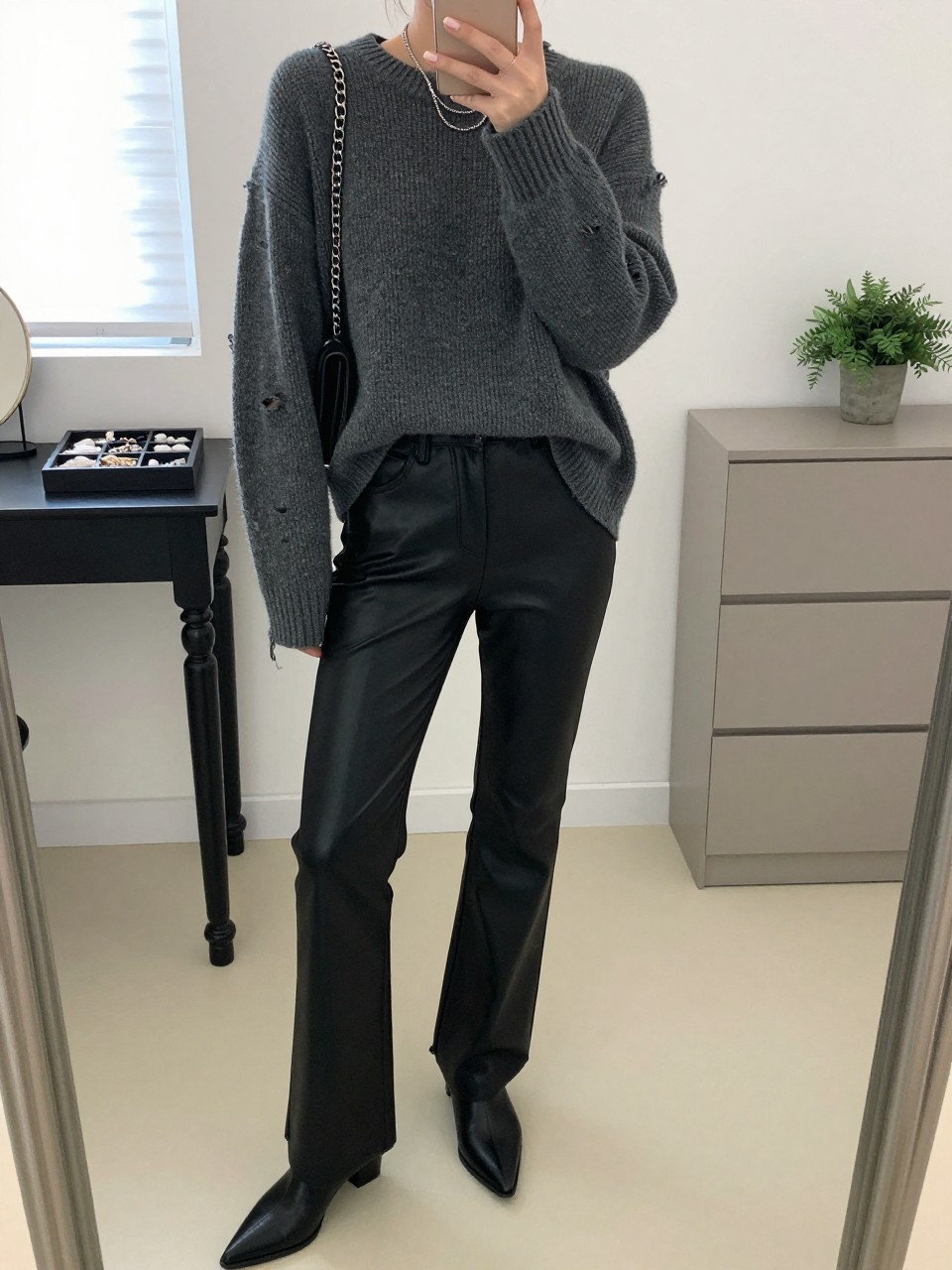Mirror selfie of a woman wearing straight-leg black faux-leather pants, an oversized charcoal-grey distressed knit sweater with small worn openings, black pointed-toe ankle boots, a silver chain shoulder bag, and layered silver necklaces, clearly showing the exact clothing items, colors, fabrics, and accessories listed, holding a phone in front of a minimal dressing room mirror, cropped at the neck so the face does not show, relaxed casual selfie pose, natural window light, a jewelry tray on the vanity and a small plant on a dresser, natural editorial setting, latest iPhone photo quality.