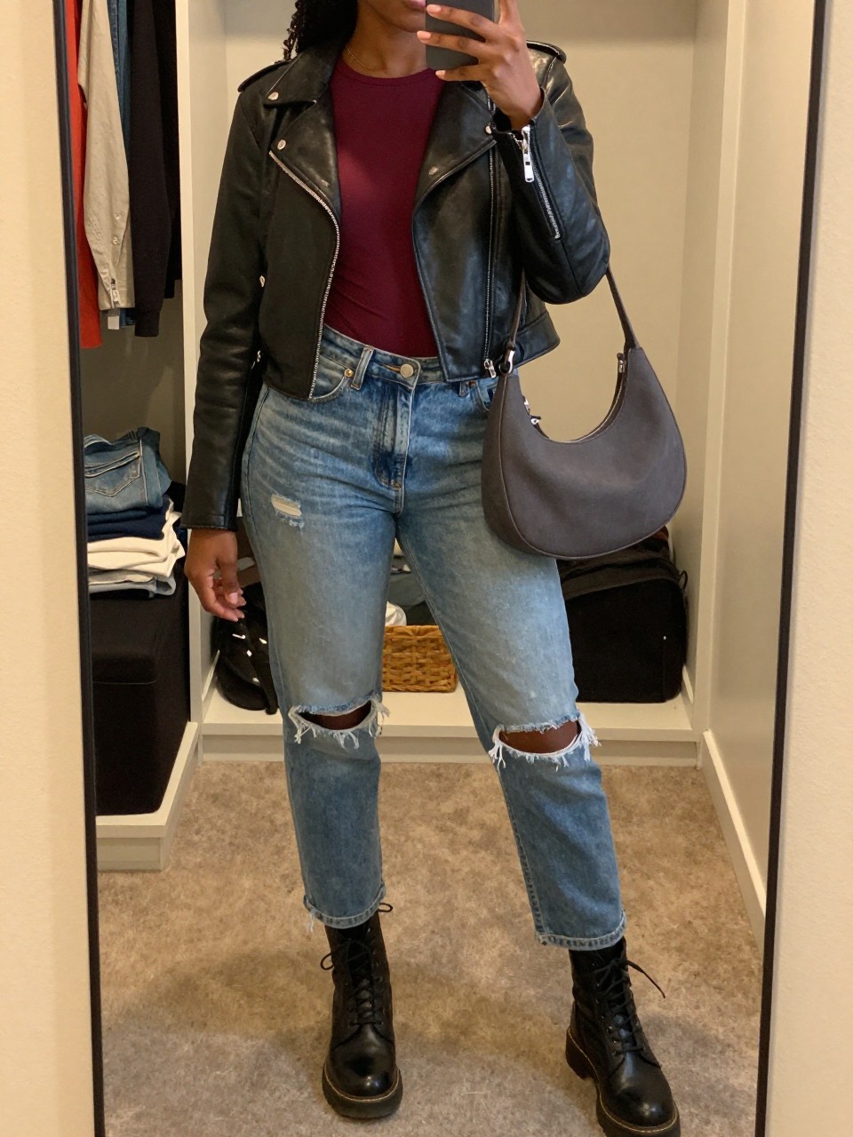 Mirror selfie of a Black woman wearing faded blue ripped mom jeans, a fitted burgundy long-sleeve top, a cropped black leather moto jacket, black lace-up combat boots, and a dark grey crescent shoulder bag, clearly showing the exact clothing items, colors, fabrics, and accessories listed, holding a phone in front of a walk-in closet mirror, cropped at the neck so the face does not show, relaxed casual selfie pose, evening golden-hour lighting, an open closet with clothes visible and folded denim jeans on a bench, believable everyday setting, latest iPhone photo quality.
