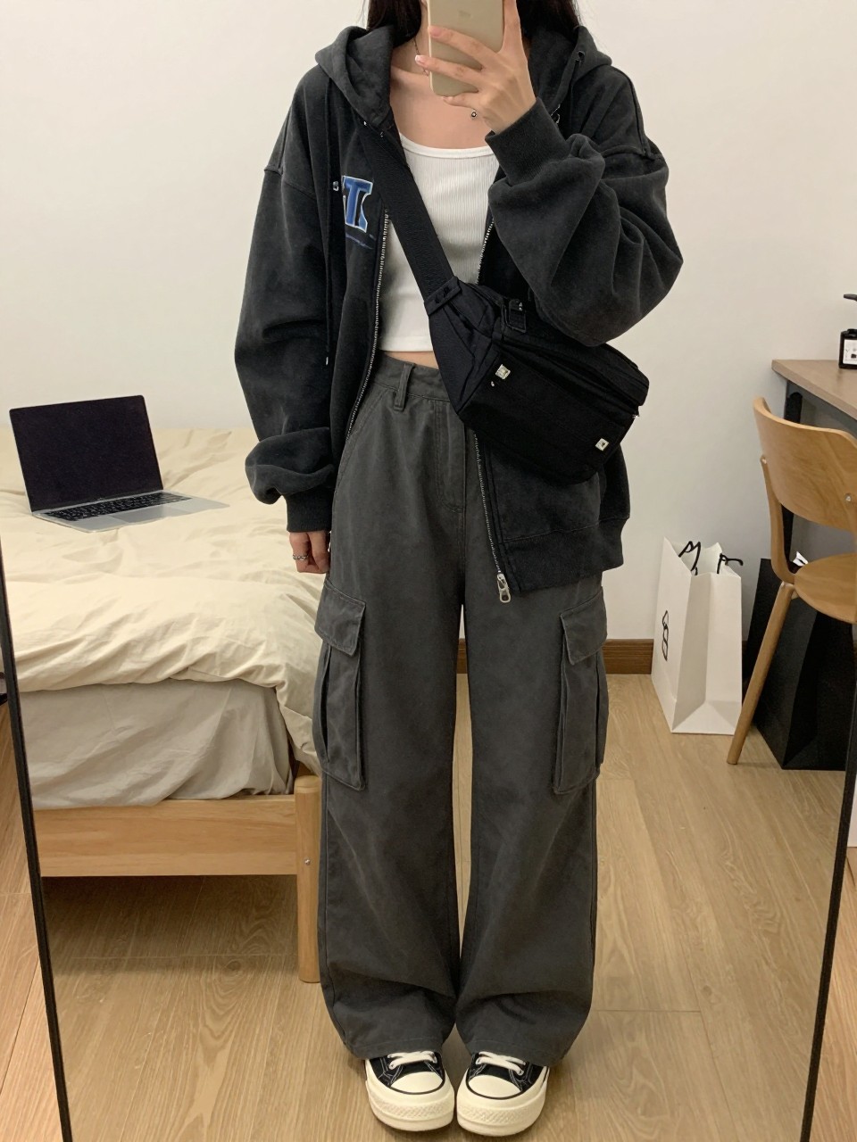 Mirror selfie of a woman wearing loose charcoal-grey cargo pants, an oversized faded black graphic hoodie, a cropped white ribbed tank top visible underneath, black and white platform sneakers, and a black nylon belt bag worn across the chest, clearly showing the exact clothing items, colors, fabrics, and accessories listed, holding a phone in front of a cozy bedroom mirror setup, cropped at the neck so the face does not show, relaxed casual selfie pose, soft indoor lighting, a laptop on the bed and shopping bags beside a chair, realistic casual atmosphere, latest iPhone photo quality.