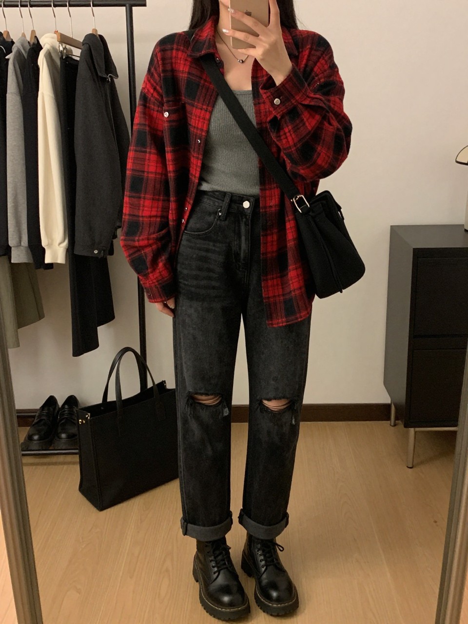 Mirror selfie of a woman wearing distressed black straight-leg jeans, an oversized red and black flannel plaid shirt worn open, a fitted charcoal-grey ribbed tank top, black leather combat boots, and a black canvas crossbody bag, clearly showing the exact clothing items, colors, fabrics, and accessories listed, holding a phone in front of a stylish city apartment mirror, cropped at the neck so the face does not show, relaxed casual selfie pose, warm moody lighting, a clothing rack with garments behind and a tote bag resting on the floor, natural and uncluttered, latest iPhone photo quality.