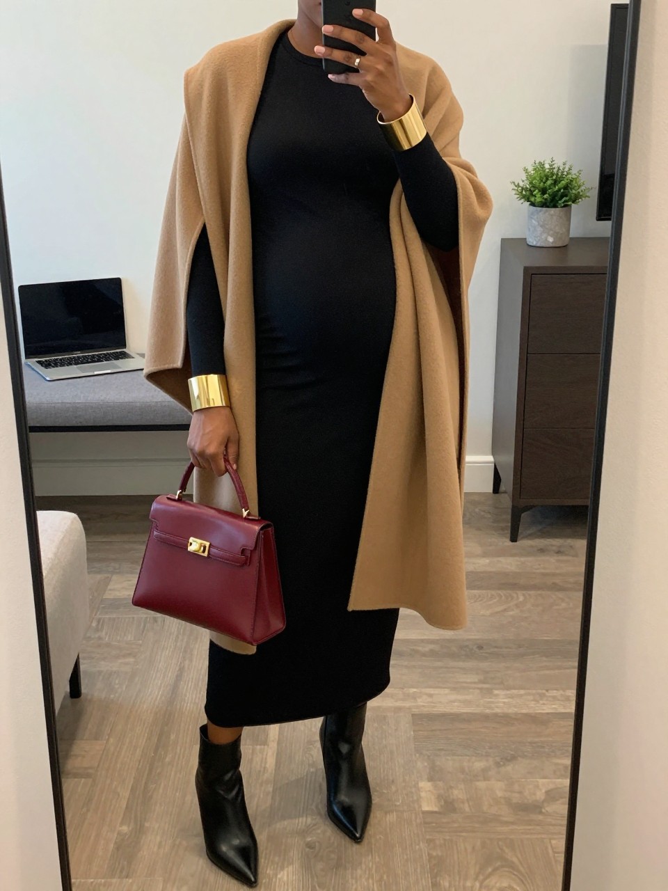 Mirror selfie of a Black pregnant woman wearing a fitted black long-sleeve maternity column maxi dress, an open camel wool cape coat, black pointed-toe ankle boots, an oxblood top-handle bag, and bold gold cuff bracelets, clearly showing the exact clothing items, colors, fabrics, and accessories listed, holding a phone in front of a stylish city apartment mirror, cropped at the neck so the face does not show, with a relaxed casual selfie pose, soft indoor lighting, a laptop on a bench and a small plant on a dresser, modern realistic setting, latest iPhone photo quality.