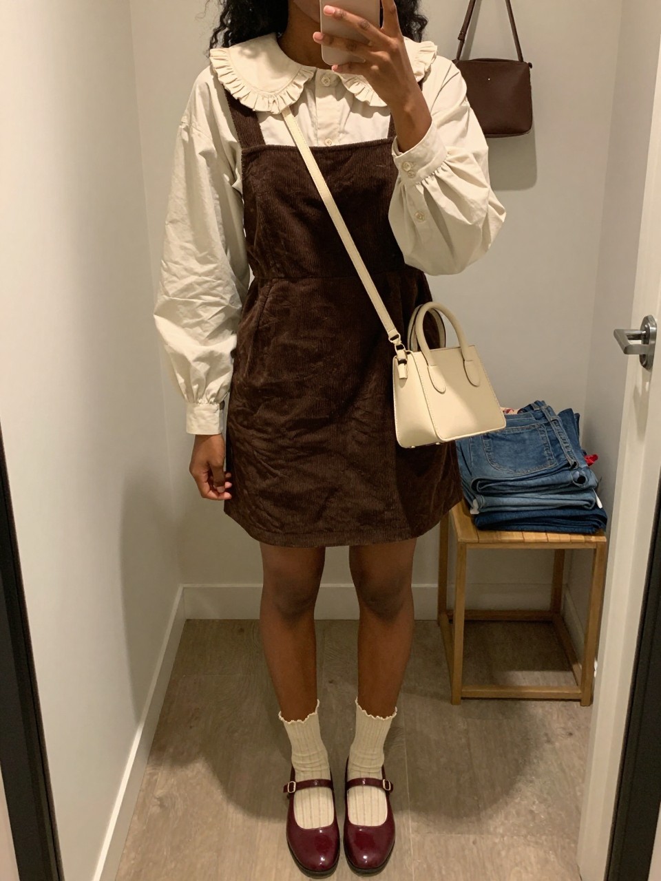 Image Prompt mirror selfie of a Black woman wearing an ivory cotton blouse with a ruffled collar and puff sleeves under a chocolate corduroy square-neck pinafore mini dress, cream ribbed ankle socks with scalloped edges visible above burgundy leather Mary Jane shoes, and a cream structured mini crossbody bag, clearly showing the exact clothing items, colors, fabrics, collar, socks, shoes, and bag listed in the Outfit Pieces section, holding a phone in front of a mirror, cropped at NECK so the face does not show, relaxed playful selfie pose in a walk-in closet mirror, warm moody lighting, with a handbag hanging on a door handle and folded denim jeans on a bench, realistic cotton, ruffles, corduroy, ribbed knit, and leather textures, natural casual mirror composition, latest iPhone photo quality