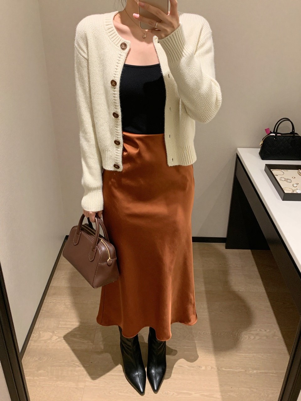 Image Prompt mirror selfie of a woman wearing a cream fitted knit cardigan with brown Western-style stitching buttoned halfway over a black fitted square-neck top tucked into a rust satin midi slip skirt, black pointed-toe ankle boots, and a chocolate structured mini handbag, clearly showing the exact clothing items, colors, stitching, fabrics, boots, and bag listed in the Outfit Pieces section, holding a phone in front of a mirror, cropped at NECK so the face does not show, relaxed polished selfie pose in a minimal dressing room, warm moody lighting, with a jewelry tray on the vanity and a cosmetic bag on the counter, realistic knit, stitching, satin, and leather textures, latest iPhone photo quality