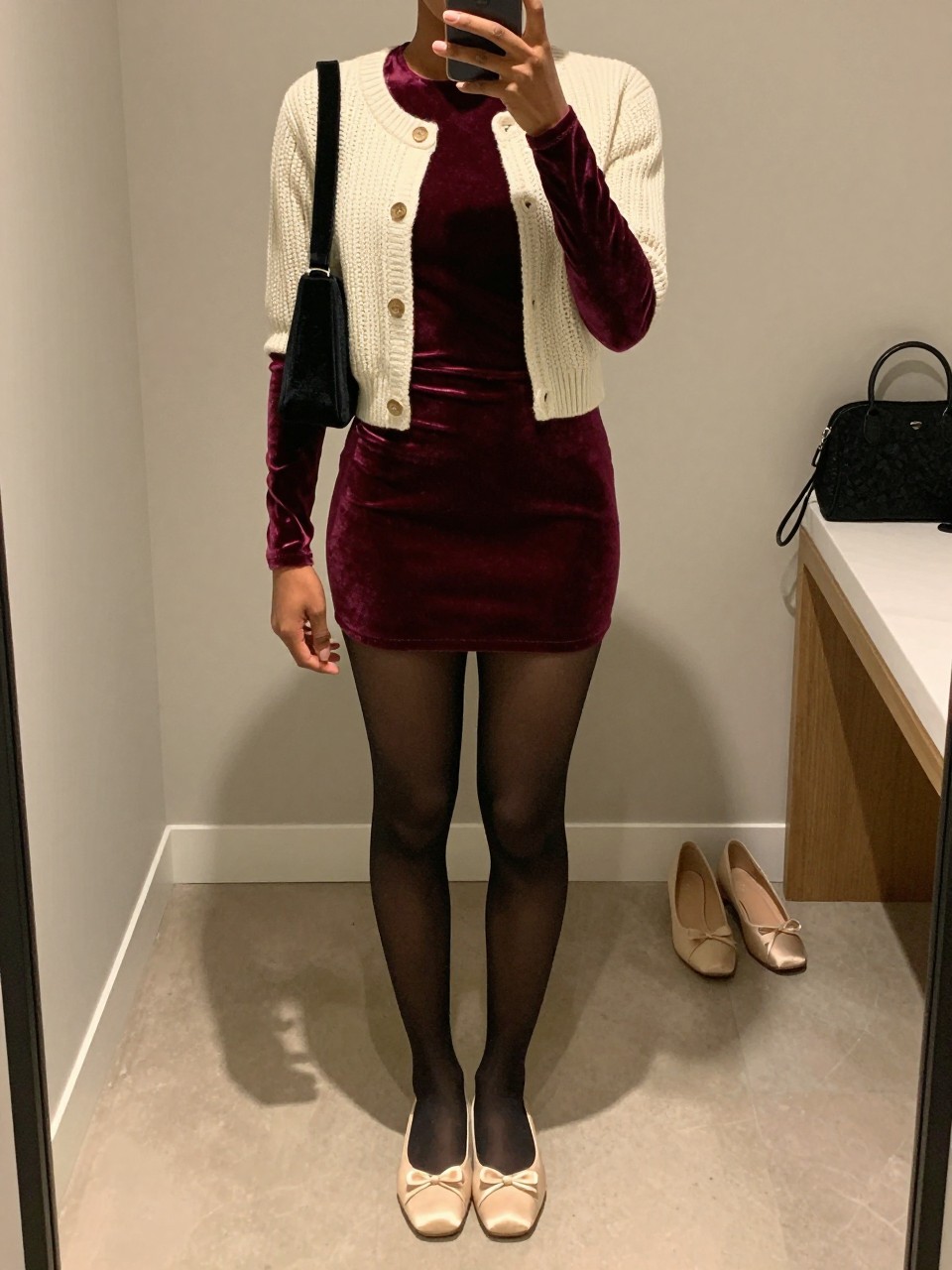 Image Prompt mirror selfie of a Black woman wearing a burgundy velvet long-sleeve mini dress, an open cream cropped fine-knit cardigan, sheer black tights, cream ballet flats with small satin bows, and a black velvet shoulder bag, clearly showing the exact clothing items, colors, fabrics, tights, bows, shoes, and bag listed in the Outfit Pieces section, holding a phone in front of a mirror, cropped at NECK so the face does not show, relaxed elegant selfie pose in a minimal dressing room, warm moody lighting, with a pair of heels on the floor nearby and a cosmetic bag on the counter, realistic velvet, knit, sheer tights, satin, and leather textures, latest iPhone photo quality