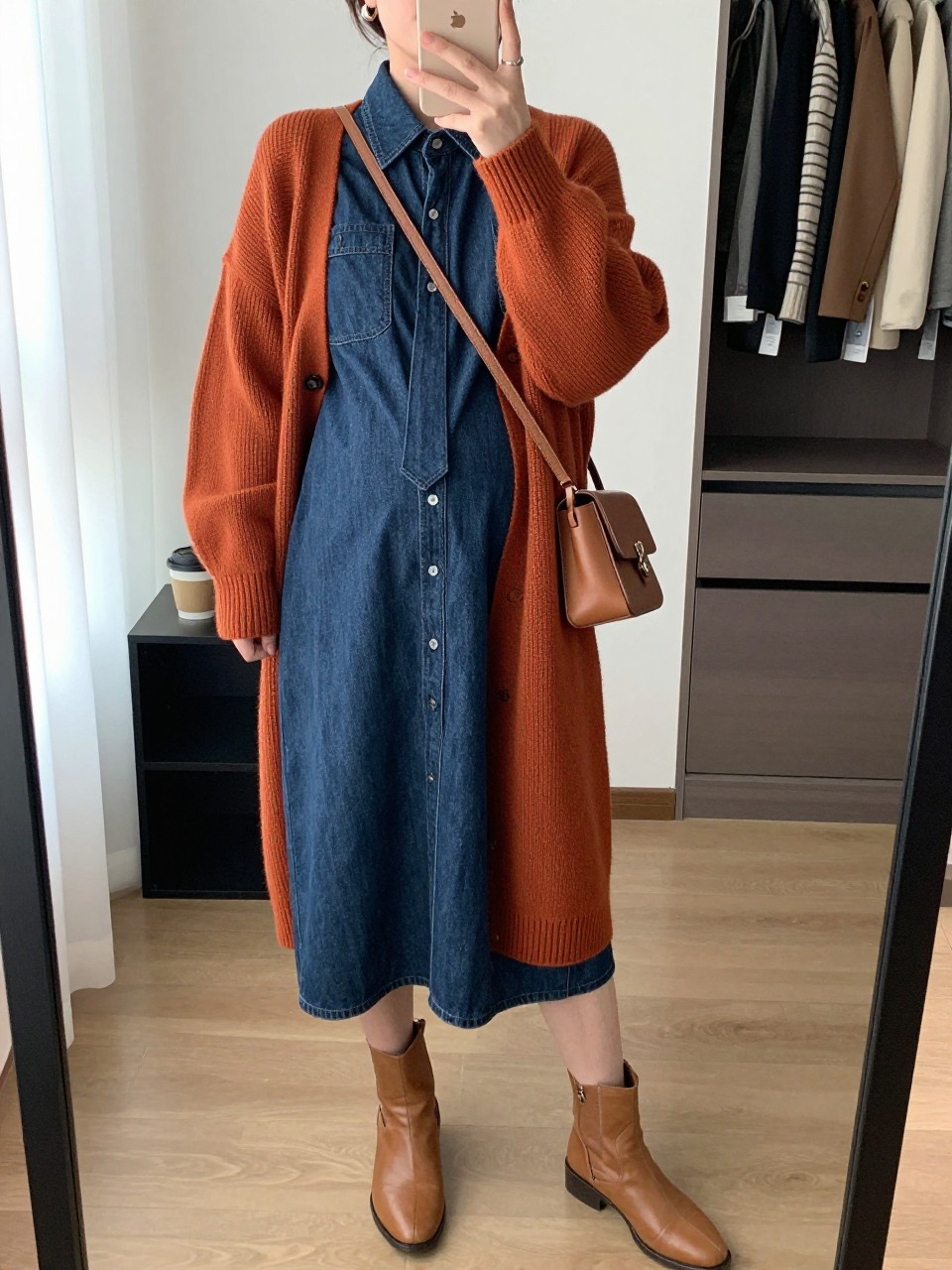 Mirror selfie of a pregnant woman wearing a long dark-wash denim maternity shirt dress with a tie positioned above the bump, an oversized rust-orange knit cardigan, tan leather ankle boots, a cognac crossbody bag, and small silver hoop earrings, clearly showing the exact clothing items, colors, fabrics, and accessories listed, holding a phone in front of a walk-in closet mirror, cropped at the neck so the face does not show, with a relaxed casual selfie pose, natural window light, an open closet with clothes visible and a coffee mug on a side table, believable everyday setting, latest iPhone photo quality.