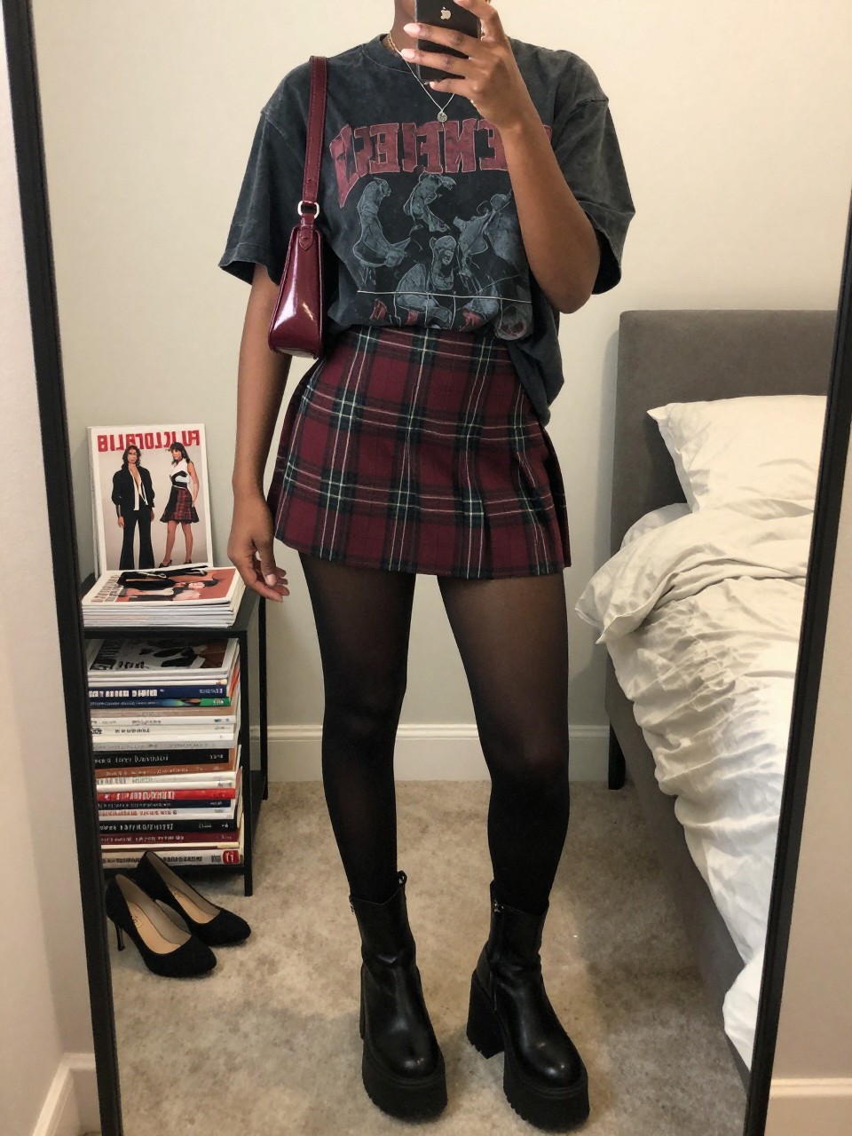 Mirror selfie of a Black woman wearing a burgundy and charcoal plaid mini skirt, a faded black oversized graphic band T-shirt, sheer black tights, chunky black platform boots, and a small burgundy shoulder bag, clearly showing the exact clothing items, colors, fabrics, and accessories listed, holding a phone in front of a feminine-themed bedroom mirror, cropped at the neck so the face does not show, relaxed casual selfie pose, soft indoor lighting, a stack of fashion magazines on a side table and a pair of heels on the floor nearby, realistic lived-in setting, latest iPhone photo quality.