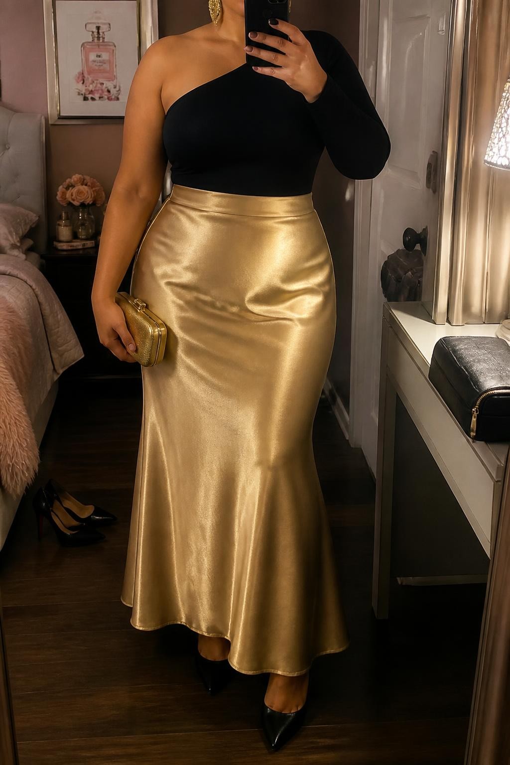 Mirror selfie of a plus size woman wearing a gold high-waisted satin maxi skirt with a softly flared shape, a fitted black one-shoulder long-sleeve top with a secure asymmetric neckline, black pointed platform heels, a small gold hard-case clutch, and chunky gold cuff earrings, clearly showing the exact clothing items, gold satin, black asymmetric top, footwear, and accessories listed, holding a phone in front of a mirror, cropped at the neck so the face is hidden, relaxed dramatic selfie pose, in a feminine-themed bedroom with warm moody lighting, a pair of heels on the floor nearby and a cosmetic bag on the counter, realistic formal evening outfit, latest iPhone photo quality.