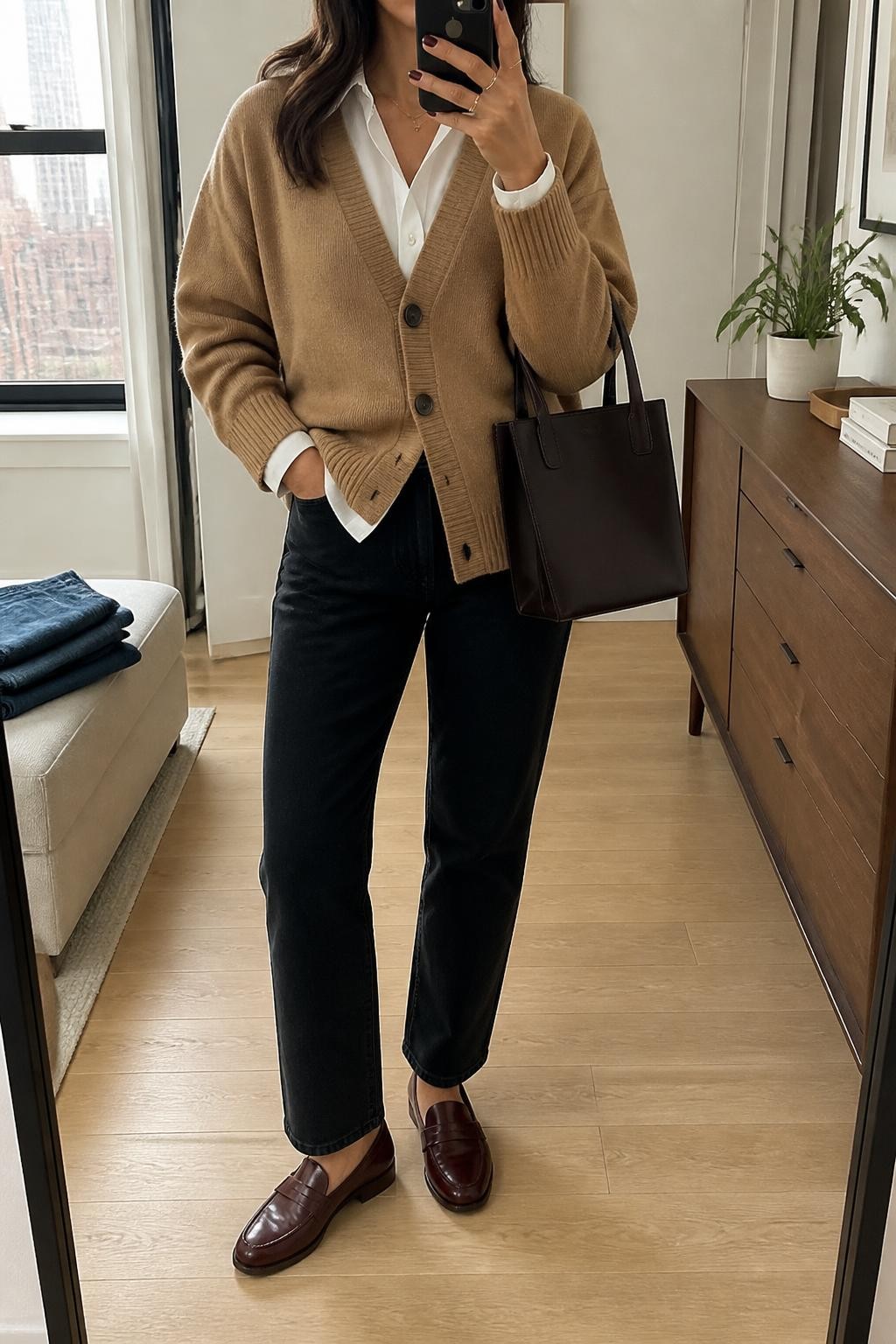 Mirror selfie of a woman wearing an oversized camel V-neck cardigan layered over a crisp white button-front shirt, black straight-leg jeans, burgundy leather loafers, and a structured dark brown handbag, clearly showing the exact clothing items, colors, fabrics, and accessories listed, holding a phone in front of a stylish city apartment mirror, cropped at the neck so the face does not show, relaxed casual selfie pose, natural window light, folded denim jeans on a bench and a small plant on a dresser, realistic and uncluttered, latest iPhone photo quality.