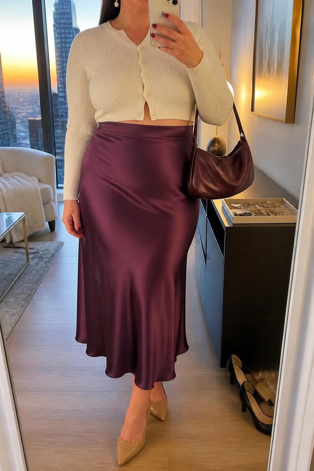 Mirror selfie of a plus size woman wearing a plum purple high-waisted satin midi skirt with a fluid bias-cut shape, a fitted ivory cropped cardigan buttoned at the front, nude pointed block heels, a plum satin shoulder bag, and pearl drop earrings, clearly showing the exact clothing items, plum satin, ivory knit, colors, footwear, and accessories listed, holding a phone in front of a mirror, cropped at the neck so the face does not show, relaxed elegant selfie pose, in a stylish city apartment with evening golden-hour lighting, a jewelry tray on the dresser and a pair of heels on the floor nearby, realistic birthday brunch outfit, latest iPhone photo quality.