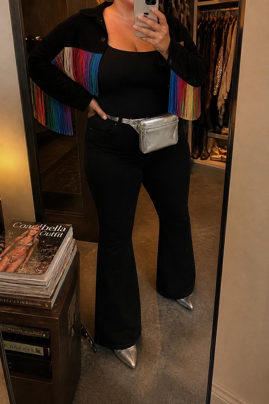 Mirror selfie of a plus size woman wearing a cropped black suede jacket with rainbow fringe across the sleeves and back, a fitted black ribbed square-neck tank top, black high-waisted flared jeans, metallic silver pointed cowboy boots, and a small silver belt bag, clearly showing the exact clothing items, rainbow fringe, black suede, black denim, silver footwear, and accessories listed, holding a phone in front of a mirror, cropped at the neck so the face is hidden, relaxed show-stopping selfie pose, in a walk-in closet mirror with warm moody lighting, an open closet with clothes visible and a stack of fashion magazines on a side table, realistic headline Coachella outfit, latest iPhone photo quality.