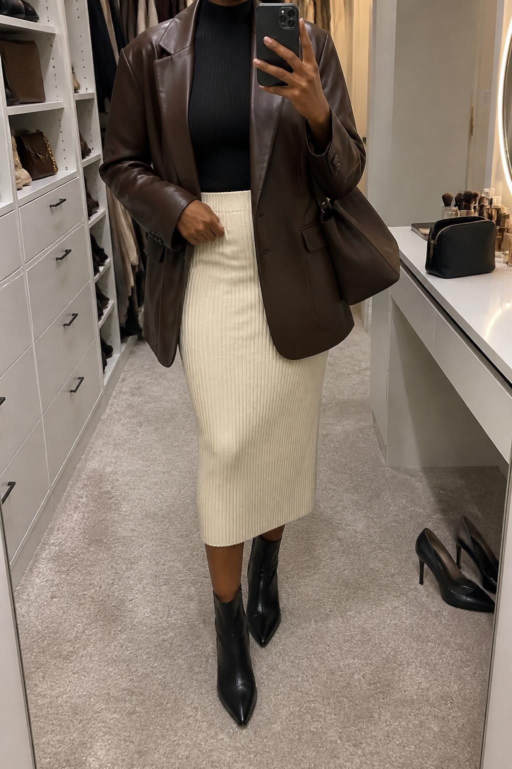 mirror selfie of a Black woman wearing an open espresso brown faux-leather blazer over a black fitted mock-neck knit top, a cream ribbed knit midi skirt with a below-the-knee hem, black pointed-toe ankle boots, and an espresso structured shoulder bag, clearly showing the exact clothing items, colors, fabrics, boots, and bag listed in the Outfit Pieces section, holding a phone in front of a mirror, cropped at NECK so the face does not show, relaxed confident selfie pose in a walk-in closet mirror, soft indoor lighting, with a pair of heels on the floor nearby and a cosmetic bag on the vanity, realistic faux-leather, ribbed knit, and leather textures, natural mirror photo, latest iPhone photo quality