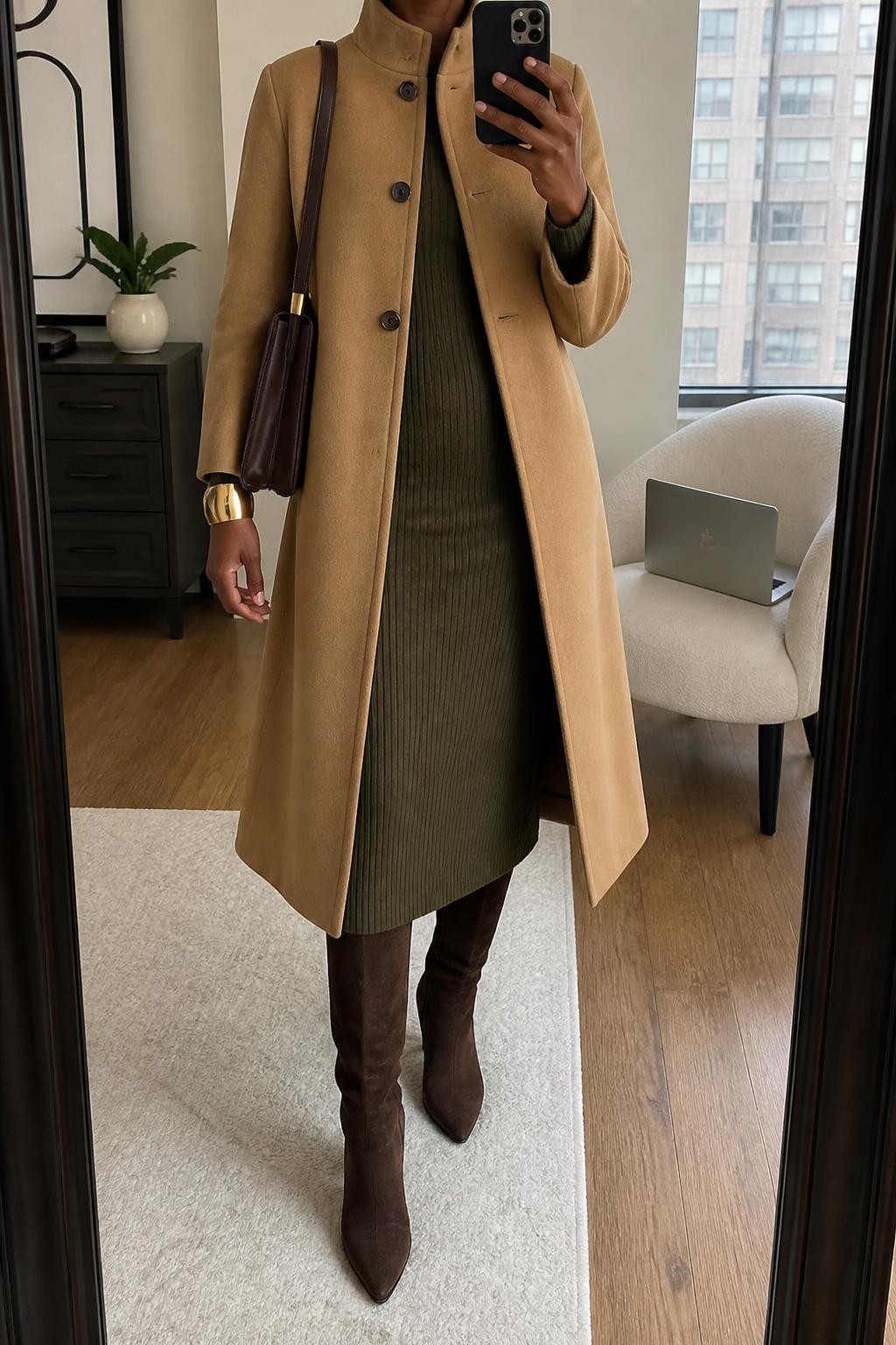 mirror selfie of a Black woman wearing a camel wool-blend funnel-neck coat fastened only through the upper section over an olive ribbed long-sleeve midi dress, chocolate suede knee-high boots, a dark brown structured shoulder bag, and a wide brushed gold cuff, clearly showing the exact clothing items, colors, fabrics, boots, bag, and bracelet listed in the Outfit Pieces section, holding a phone in front of a mirror, cropped at NECK so the face does not show, relaxed professional selfie pose in a stylish city apartment, soft indoor lighting, with a laptop resting on a chair and a small plant on a dresser, realistic wool, ribbed knit, suede, and leather textures, latest iPhone photo quality