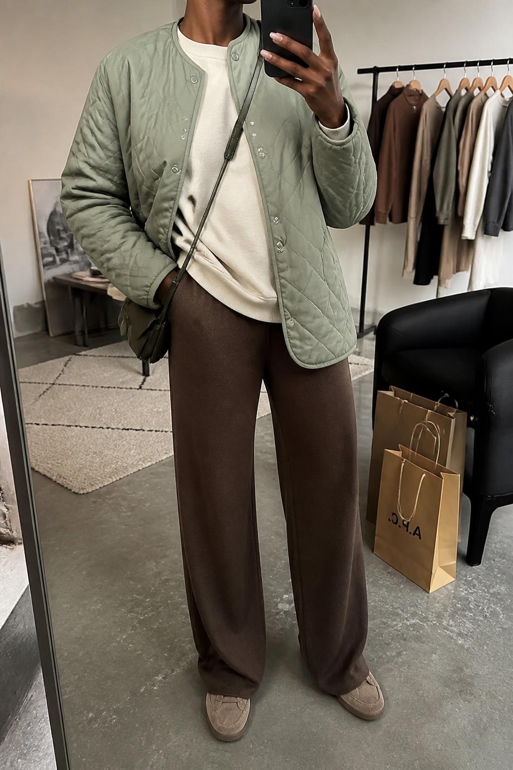 mirror selfie of a Black woman wearing an open muted sage diamond-quilted jacket over a cream brushed-cotton crewneck sweatshirt falling naturally over chocolate wide-leg knit trousers, taupe suede sneakers, and a dark olive nylon crossbody bag, clearly showing the exact clothing items, colors, fabrics, sneakers, and bag listed in the Outfit Pieces section, holding a phone in front of a mirror, cropped at NECK so the face does not show, relaxed casual selfie pose in a fashion studio mirror corner, soft indoor lighting, with a clothing rack holding a few garments behind her and shopping bags beside a chair, realistic quilting, brushed cotton, knit, suede, and nylon textures, latest iPhone photo quality