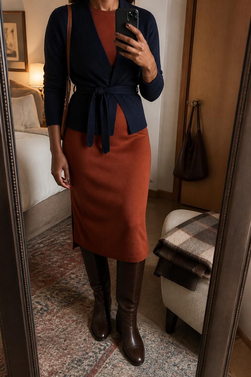 mirror selfie of a Black woman wearing a navy fine-knit wrap cardigan tied slightly above the natural waist with its matching navy knit waist tie over a rust short-sleeve knit midi dress, dark brown leather knee-high boots, and a cognac structured shoulder bag, clearly showing the exact clothing items, colors, fabrics, belt, boots, and bag listed in the Outfit Pieces section, holding a phone in front of a mirror, cropped at NECK so the face does not show, relaxed professional selfie pose in a cozy bedroom mirror setup, warm moody lighting, with a folded blanket on a chair and a handbag hanging on a door handle, realistic knit and leather textures, natural fabric folds, latest iPhone photo quality