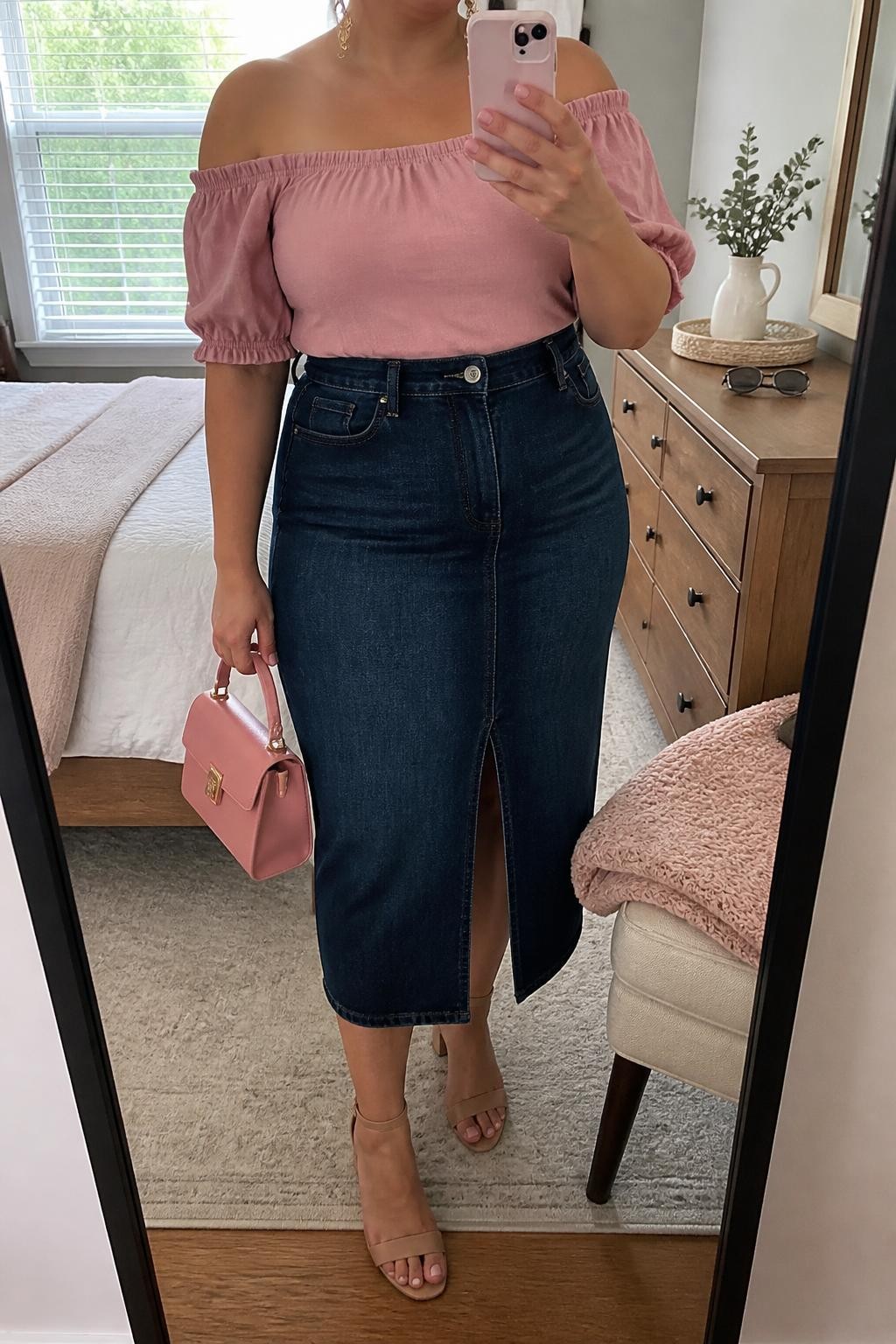 Mirror selfie of a plus size woman wearing a dark indigo high-waisted denim midi skirt with a front slit, a soft pink off-shoulder blouse with gathered short sleeves, nude block-heel sandals, a structured pink mini handbag, and delicate gold drop earrings, clearly showing the exact clothing items, dark denim, pink blouse, sleeve details, footwear, and accessories listed, holding a phone in front of a mirror, cropped at the neck so the face does not appear, relaxed feminine selfie pose, in a cozy bedroom mirror setup with natural window light, a folded blanket on a chair and sunglasses placed on a dresser, realistic casual brunch outfit, latest iPhone photo quality.