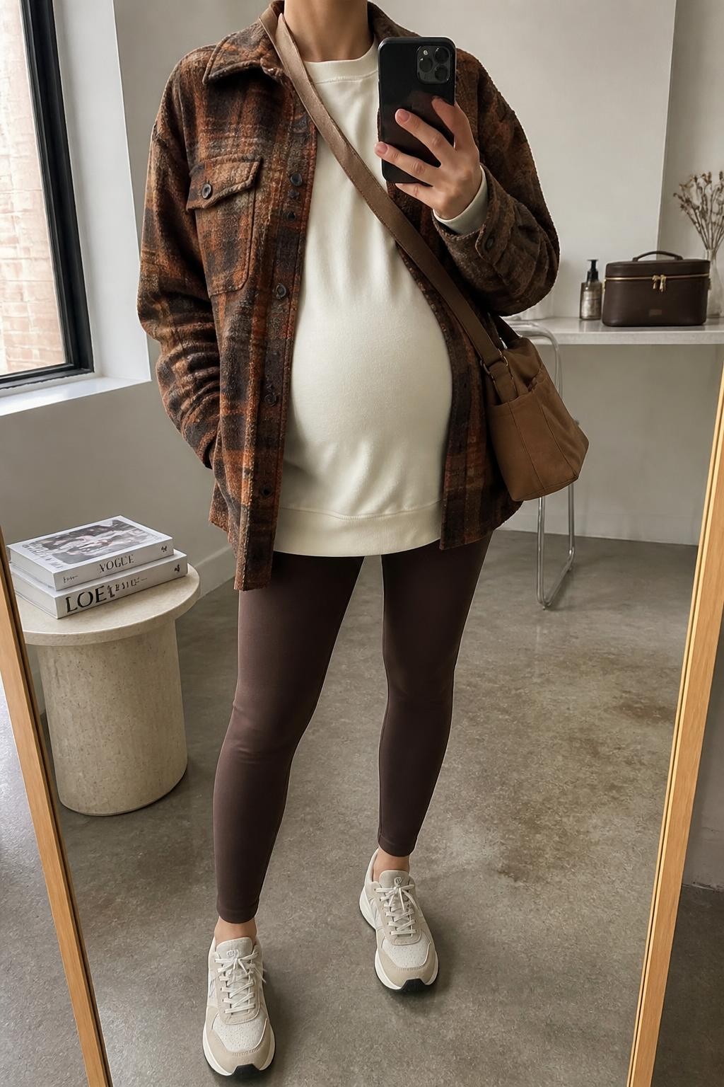 mirror selfie of a pregnant woman wearing an open brown and rust plaid wool-blend shacket over an ivory longline maternity sweatshirt falling below the jacket, chocolate over-the-bump maternity leggings, beige cushioned sneakers, and a brown canvas crossbody bag resting beside the visible baby bump, clearly showing the exact clothing items, colors, plaid pattern, fabrics, sneakers, and bag listed in the Outfit Pieces section, holding a phone in front of a mirror, cropped at NECK so the face does not show, relaxed casual maternity selfie pose in a fashion studio mirror corner, natural window light, with a stack of fashion magazines on a side table and a cosmetic bag on the counter, realistic wool, sweatshirt fabric, stretch leggings, canvas, and sneaker textures, latest iPhone photo quality