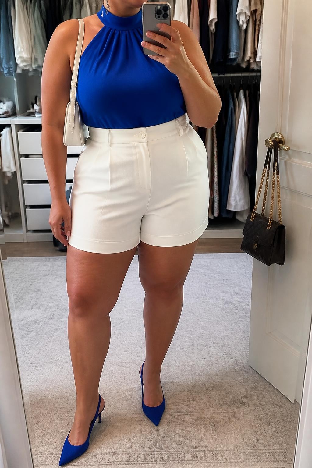 Mirror selfie of a plus size woman wearing crisp white high-waisted tailored shorts with relaxed thigh openings, a cobalt blue halter-neck top with a wide supportive neck band, cobalt pointed slingback heels, a small white leather shoulder bag, and silver drop earrings, clearly showing the exact clothing items, white tailoring, cobalt top, colors, footwear, and accessories listed, holding a phone in front of a mirror, cropped at the neck so her face does not appear, relaxed confident selfie stance, in a walk-in closet mirror with soft indoor lighting, an open closet with clothes visible and a handbag hanging on a door handle, realistic rooftop brunch styling, latest iPhone photo quality.