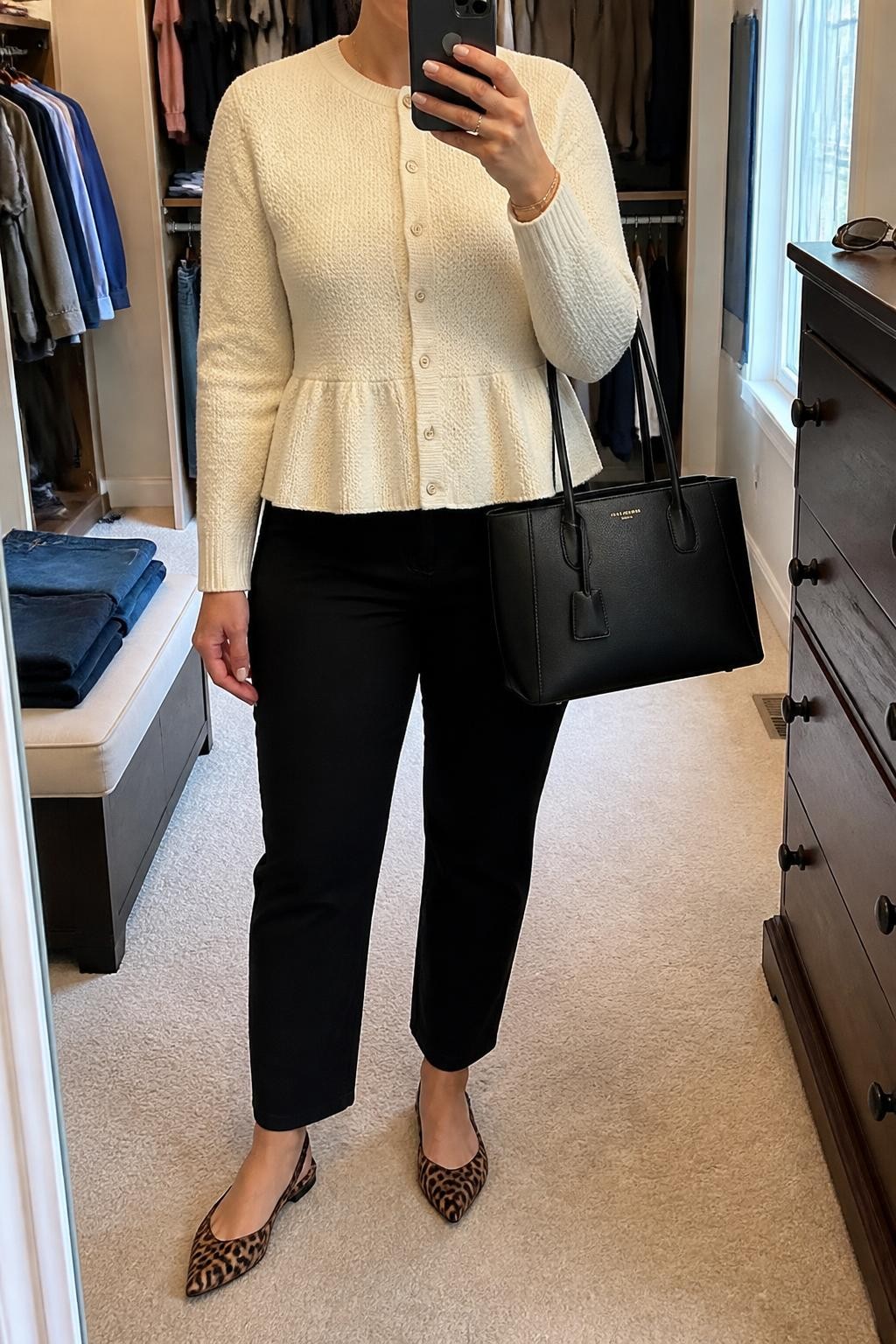 mirror selfie of a midsize woman wearing a fully buttoned cream textured peplum cardigan, black cigarette jeans, leopard-print slingback flats, and a black structured handbag carried at her side, clearly showing the exact clothing items, colors, textures, shoes, and handbag listed in the Outfit Pieces section, holding a phone in front of a mirror, cropped at NECK so the face does not show, relaxed casual selfie pose in a walk-in closet mirror, natural window light, with folded denim jeans on a bench and sunglasses placed on a dresser, realistic knit, denim, and printed shoe textures, unstaged home setting, latest iPhone photo quality