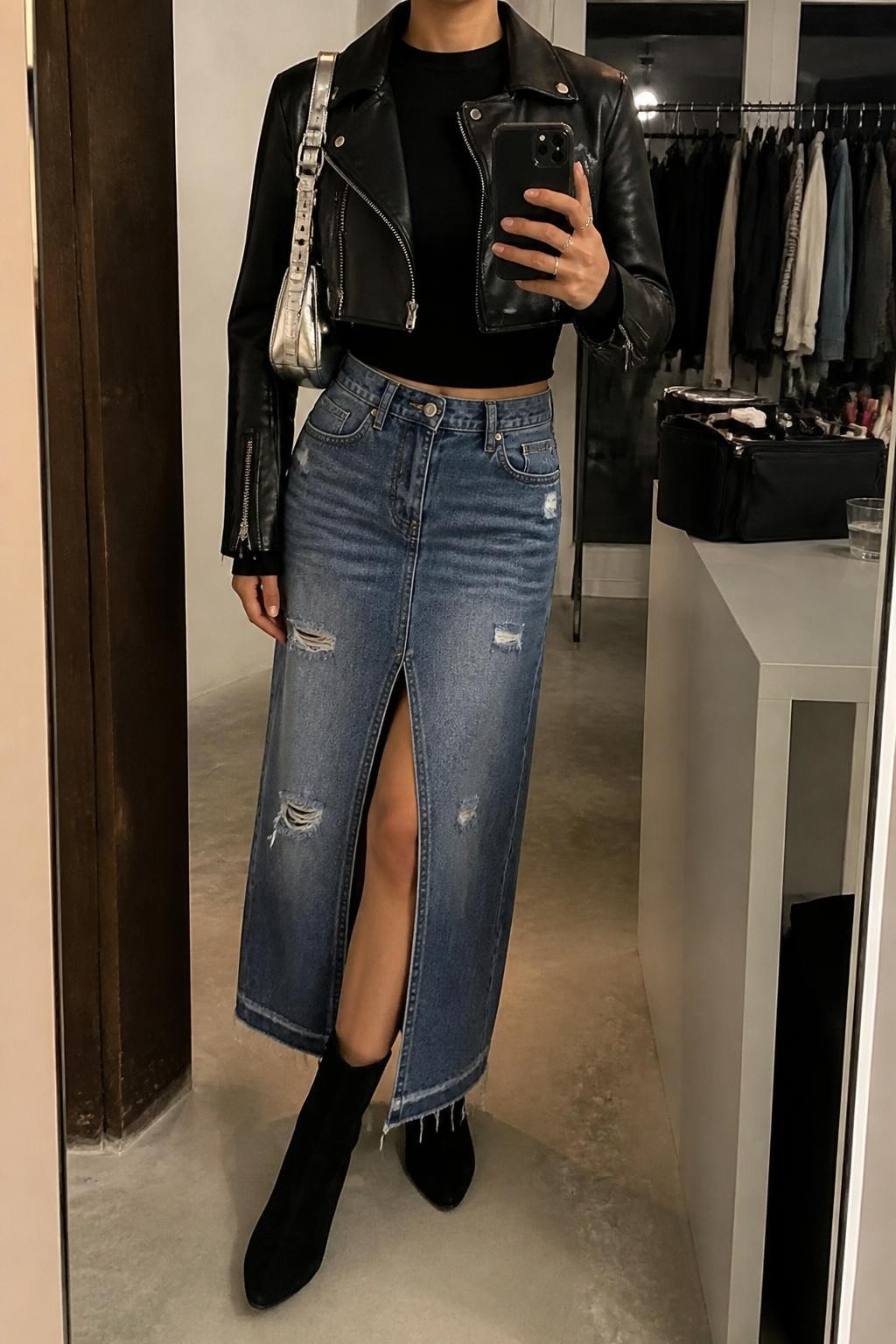 Mirror selfie of a woman wearing a distressed medium-blue denim maxi skirt with a thigh-high front slit, fitted black long-sleeve top, cropped black leather moto jacket, black suede ankle boots, and a metallic silver shoulder bag, clearly showing the exact clothing items, colors, fabrics, and accessories listed, holding a phone in front of a fashion studio mirror corner, cropped at the neck so the face does not show, relaxed casual selfie pose, warm moody lighting, a clothing rack with garments behind and a makeup bag on the counter, natural editorial setting, latest iPhone photo quality.