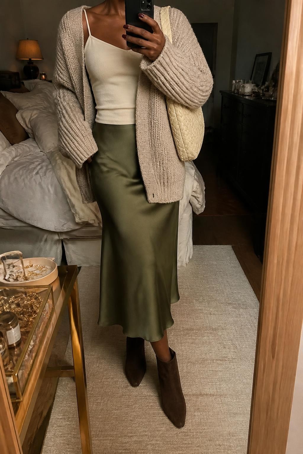Mirror selfie of a Black woman wearing an oversized oatmeal chunky-knit cardigan over a fitted cream camisole, an olive-green satin midi skirt, dark brown suede ankle boots, and a cream woven shoulder bag, clearly showing the exact clothing items, colors, fabrics, and accessories listed, holding a phone in front of a cozy bedroom mirror setup, cropped at the neck so the face does not show, relaxed casual selfie pose, warm moody lighting, soft pillows on an unmade bed and a jewelry tray on the vanity, realistic lived-in setting, latest iPhone photo quality.