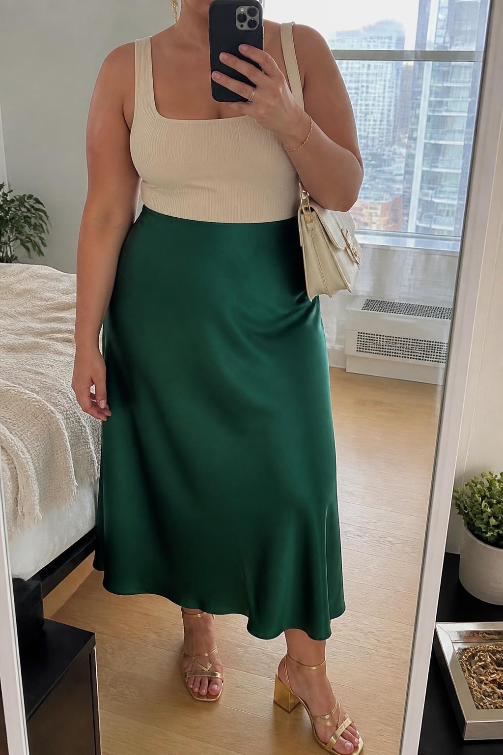 Mirror selfie of a plus size woman wearing an emerald green high-waisted satin midi skirt with a fluid bias-cut shape, a fitted cream ribbed square-neck bodysuit, metallic gold block-heel sandals, a structured cream shoulder bag, and delicate gold drop earrings, clearly showing the exact clothing items, emerald satin, cream ribbed fabric, colors, footwear, and accessories listed, holding a phone in front of a mirror, cropped at the neck so the face does not show, relaxed elegant selfie pose, in a stylish city apartment with natural window light, a jewelry tray on the dresser and a small plant nearby, natural unstaged appearance, latest iPhone photo quality.