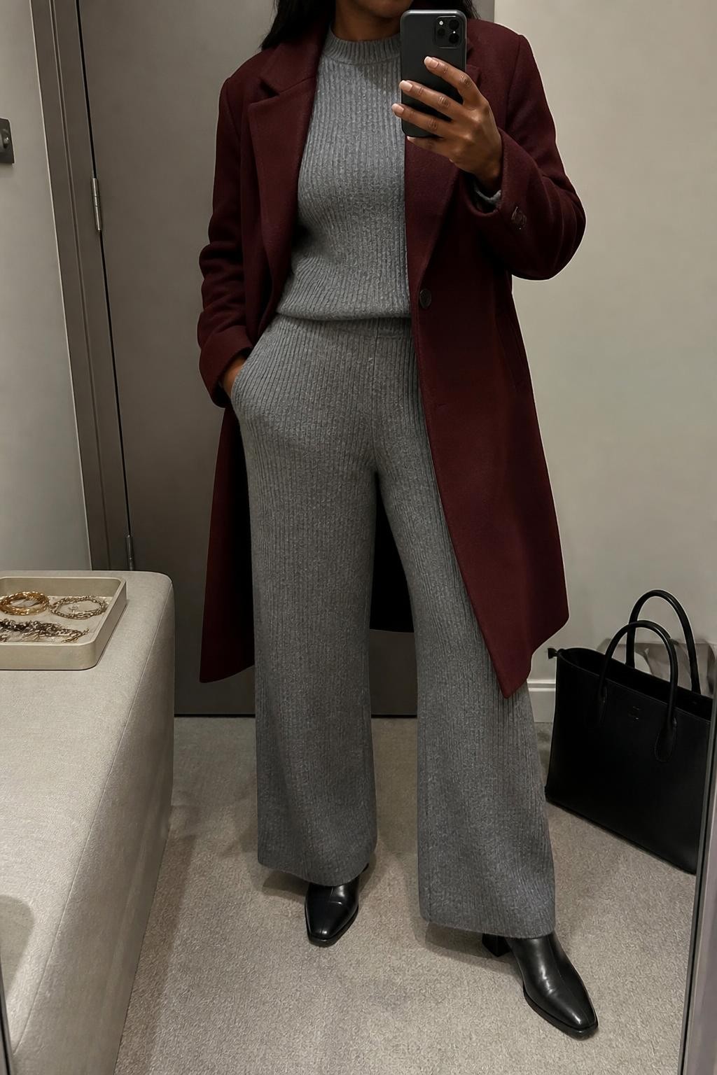 mirror selfie of a Black woman wearing an open burgundy single-breasted wool-blend coat over a matching heather gray ribbed crewneck sweater and heather gray ribbed wide-leg knit trousers, black leather ankle boots, and a black structured travel tote, clearly showing the exact clothing items, colors, fabrics, boots, and bag listed in the Outfit Pieces section, holding a phone in front of a mirror, cropped at NECK so the face does not show, relaxed travel-ready selfie pose in a minimal dressing room, soft indoor lighting, with a tote bag resting on the floor and a jewelry tray on a nearby surface, realistic wool, ribbed knit, and leather textures, latest iPhone photo quality