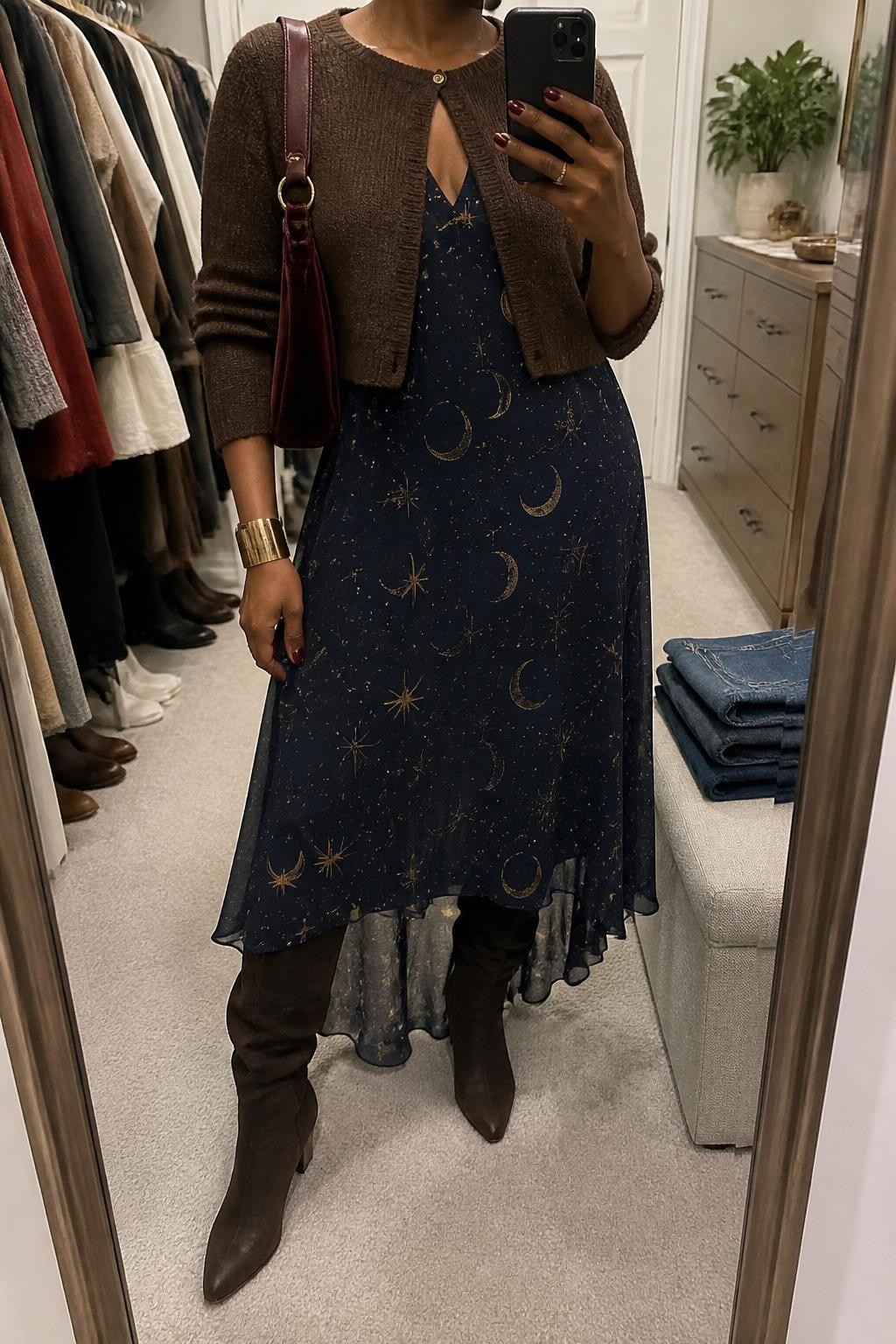 mirror selfie of a Black woman wearing a navy chiffon maxi dress with a clear gold moon and star print, a chocolate cropped knit cardigan buttoned only at the top, chocolate suede knee-high boots, a burgundy velvet shoulder bag, and an antique gold cuff bracelet, clearly showing the exact clothing items, colors, celestial print, fabrics, boots, bag, and bracelet listed in the Outfit Pieces section, holding a phone in front of a mirror, cropped at NECK so the face does not show, relaxed feminine selfie pose in a walk-in closet mirror, soft indoor lighting, with folded denim jeans on a bench and a small plant on a dresser, realistic chiffon, knit, suede, and velvet textures, latest iPhone photo quality