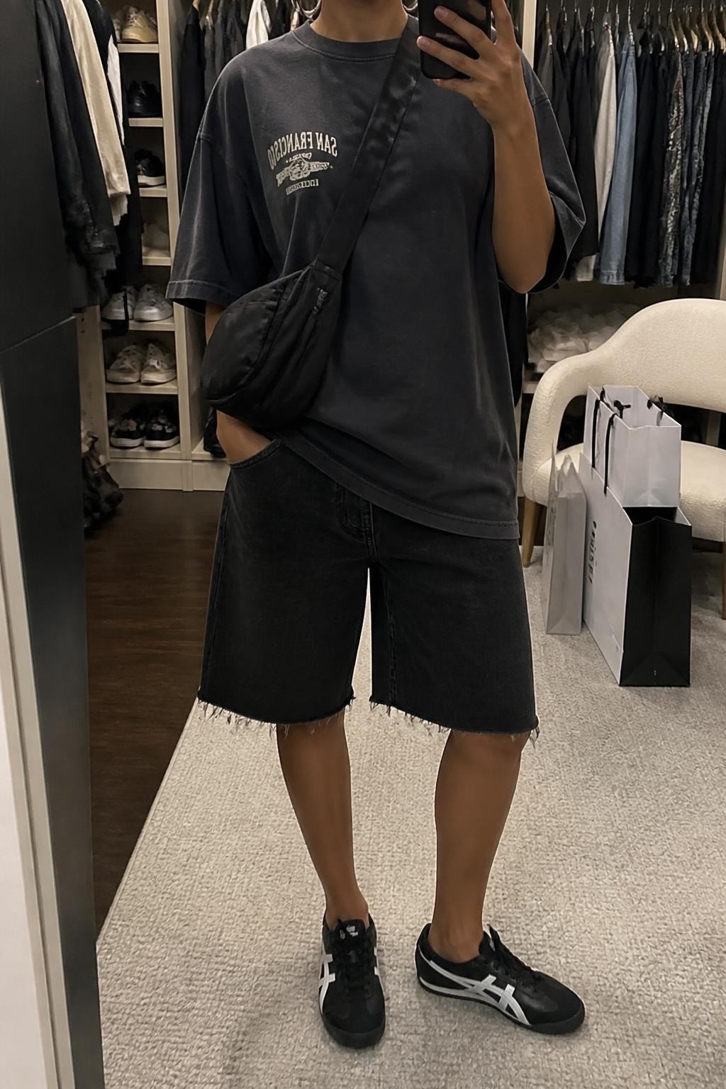 mirror selfie of a woman with a medium brown skin tone wearing loose raw-hem black denim jorts, an oversized charcoal cotton T-shirt with a small faded cream graphic, black and white sporty low-profile sneakers, a black nylon crossbody bag and chunky silver hoop earrings, exact clothing and accessories listed with nothing additional, holding a phone in front of a mirror, cropped at NECK so the face does not show, relaxed casual selfie pose, in a walk-in closet mirror, soft indoor lighting, open closet with clothes visible and shopping bags beside a chair, natural slightly lived-in setting, latest iPhone photo quality.