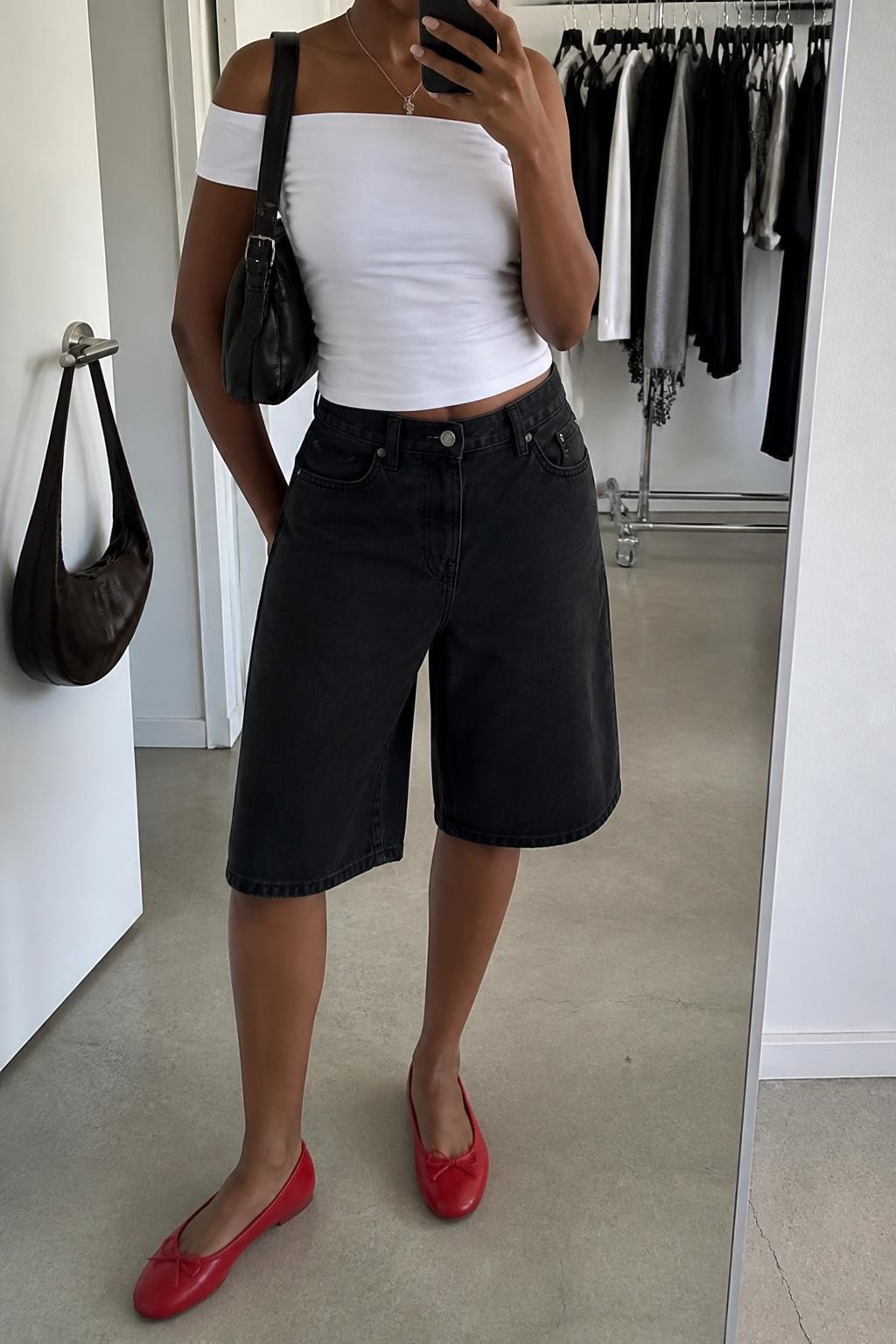 mirror selfie of a Black woman wearing oversized loose-fit black denim jorts, a fitted white stretch cotton off-shoulder top, simple rounded-toe red ballet flats, a soft black leather shoulder bag and a thin silver pendant necklace, wearing exactly the outfit described without additional layers or accessories, holding a phone in front of a mirror, cropped at NECK so the face does not show, relaxed casual selfie pose, in a fashion studio mirror corner, soft indoor lighting, clothing rack with garments behind and handbag hanging on a door handle, realistic latest iPhone photo quality.