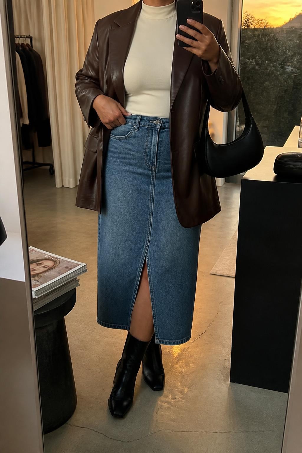 mirror selfie of a midsize woman wearing an open chocolate brown faux-leather blazer over an ivory fitted mock-neck top, a medium-wash front-slit denim midi skirt, black heeled ankle boots, and a black crescent shoulder bag, clearly showing the exact clothing items, colors, fabrics, boots, and bag listed in the Outfit Pieces section, holding a phone in front of a mirror, cropped at NECK so the face does not show, relaxed confident selfie pose in a fashion studio mirror corner, evening golden hour light, with a stack of fashion magazines on a side table and a cosmetic bag on the counter, realistic faux-leather and denim textures, slightly imperfect casual framing, latest iPhone photo quality