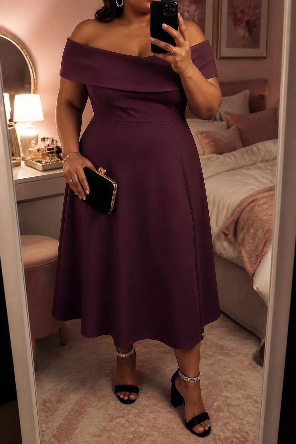 Mirror selfie of a plus size woman wearing a plum purple off-shoulder midi dress with a folded neckline, fitted waist, and softly flared skirt, black block heels with crystal ankle straps, a small black velvet clutch, and silver crystal hoop earrings, clearly showing the exact dress shape, plum color, neckline, fabrics, footwear, and accessories listed, holding a phone in front of a mirror, cropped at the neck so her face does not show, relaxed feminine selfie pose, in a feminine-themed bedroom with evening golden-hour lighting, soft pillows on an unmade bed and a jewelry tray on the vanity, realistic evening wedding outfit, latest iPhone photo quality.