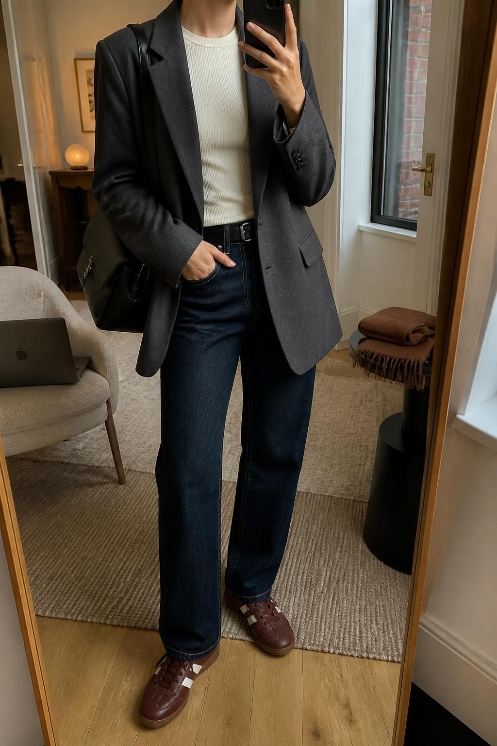 mirror selfie of a woman wearing an open charcoal relaxed-fit wool-blend blazer over a cream fitted ribbed crewneck top, dark indigo straight-leg jeans, burgundy leather sneakers, and a black structured backpack with simple hardware and no text, clearly showing the exact clothing items, colors, fabrics, sneakers, and backpack listed in the Outfit Pieces section, holding a phone in front of a mirror, cropped at NECK so the face does not show, relaxed city-ready selfie pose in a stylish city apartment, warm moody lighting, with a laptop resting on a chair and a folded scarf on a side table, realistic wool, ribbed knit, denim, and leather textures, latest iPhone photo quality