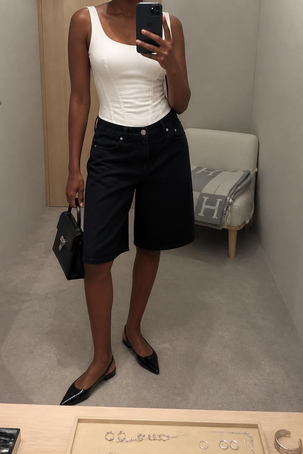 mirror selfie of a Black woman wearing loose knee-length black denim jorts with clean hems, a fitted white cotton corset-inspired top with visible seam detailing, pointed black slingback flats, a structured black mini handbag and clean silver stud earrings, wearing the exact listed outfit and no unlisted clothing pieces, holding a phone in front of a mirror, cropped at NECK so the face does not show, relaxed selfie pose, in a minimal dressing room, soft indoor lighting, jewelry tray on the vanity and folded blanket on a chair, natural realistic textures and latest iPhone photo quality.
