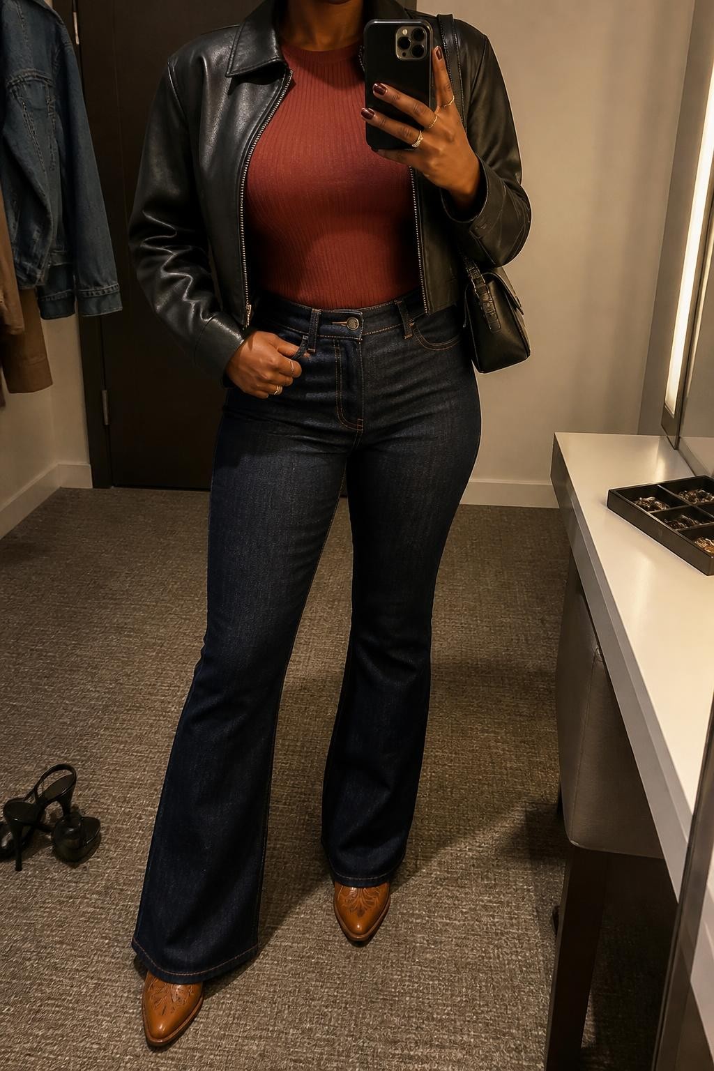 mirror selfie of a Black woman wearing an open black cropped faux-leather jacket over a rust fitted ribbed crewneck top, dark-wash high-rise flare jeans lightly skimming tan leather cowboy boots with dark brown stitching while leaving the pointed toes visible, and a black structured shoulder bag, clearly showing the exact clothing items, colors, fabrics, boots, stitching, and bag listed in the Outfit Pieces section, holding a phone in front of a mirror, cropped at NECK so the face does not show, relaxed confident selfie pose in a minimal dressing room, warm moody lighting, with a pair of heels on the floor nearby and a jewelry tray on the vanity, realistic faux-leather, ribbed knit, denim, and leather textures, latest iPhone photo quality