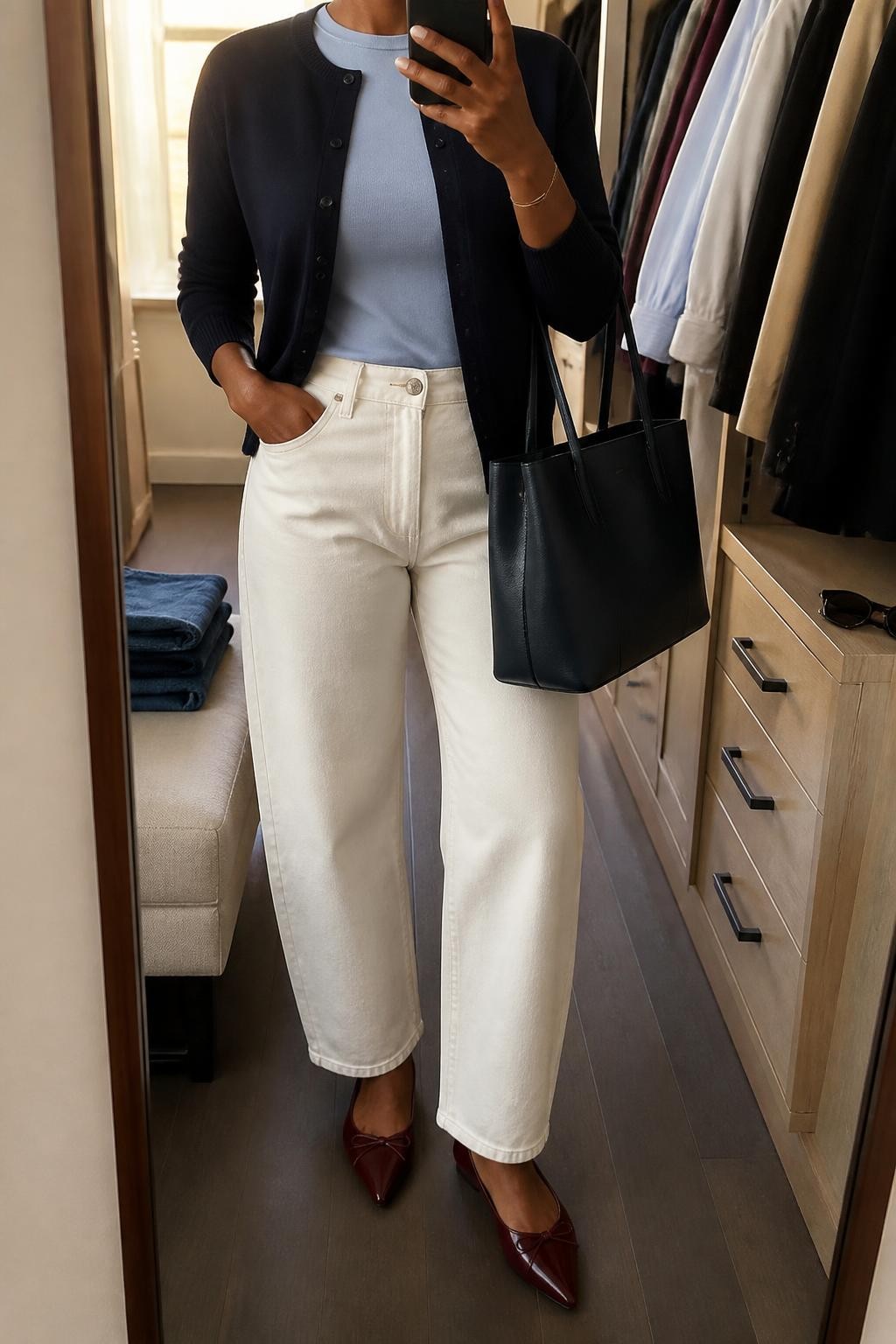 mirror selfie of a Black woman wearing a navy fine-knit cardigan buttoned halfway over a pale blue fitted crewneck top, ecru high-rise barrel-leg jeans, burgundy pointed-toe ballet flats, and a navy structured tote bag, clearly showing the exact clothing items, colors, fabrics, shoes, and bag listed in the Outfit Pieces section, holding a phone in front of a mirror, cropped at NECK so the face does not show, relaxed polished selfie pose in a walk-in closet mirror, evening golden hour light, with folded denim jeans on a bench and sunglasses placed on a dresser, realistic fine knit, cotton, denim, and leather textures, natural unstaged mirror photo, latest iPhone photo quality